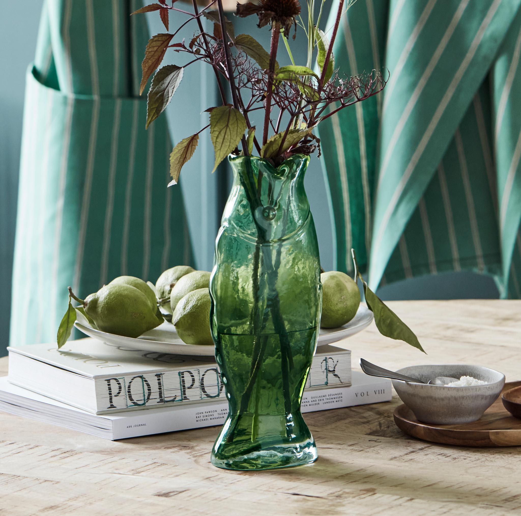 A green fish-shaped vase with a dark floral arrangement sits on a wooden table next to green fruit, red onions, and books, with striped aprons in the background.
