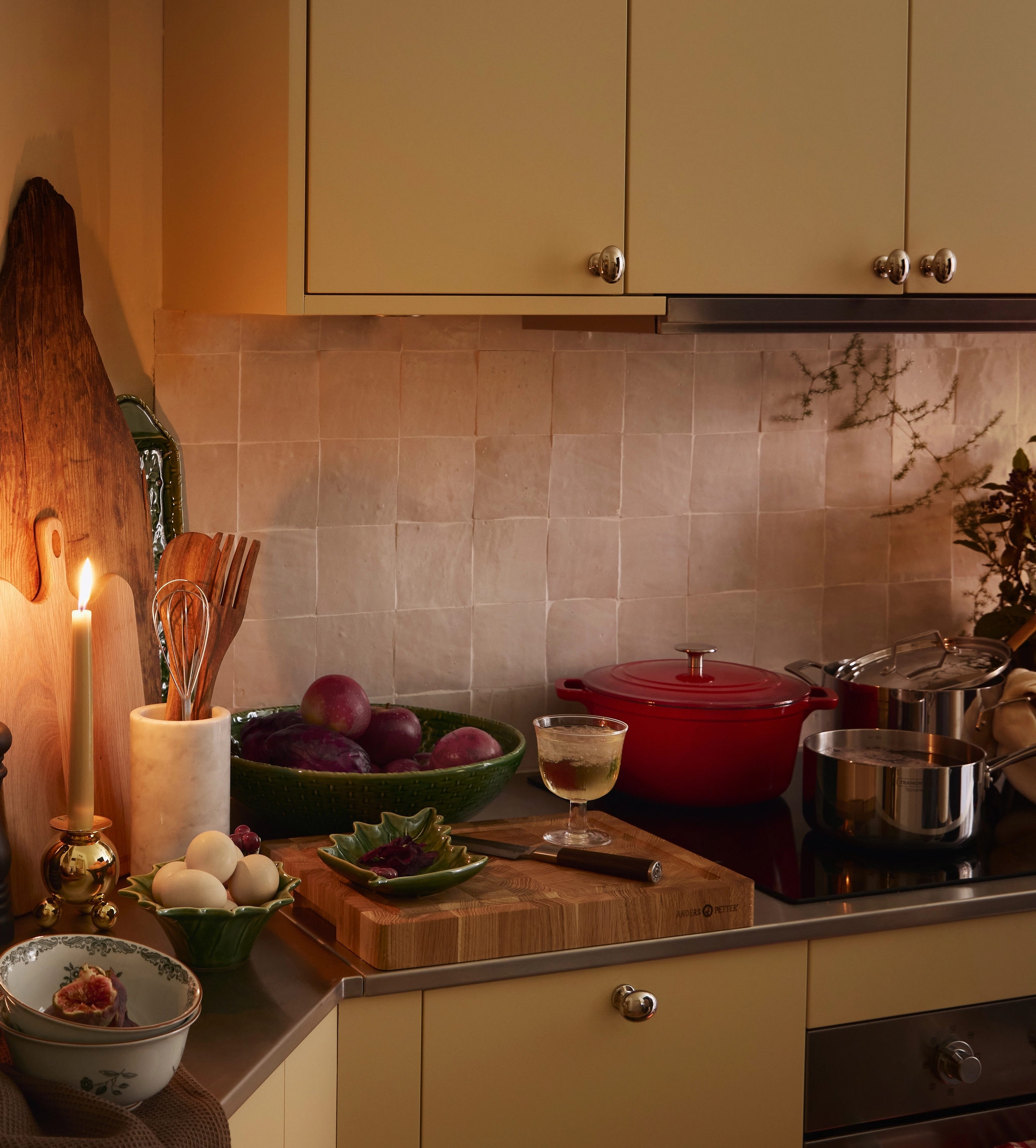 Viihtyisä keittiöntaso yöllä, yhden kynttilän valaisemana hedelmien, munien, keittiövälineiden ja mausteiden keskellä.