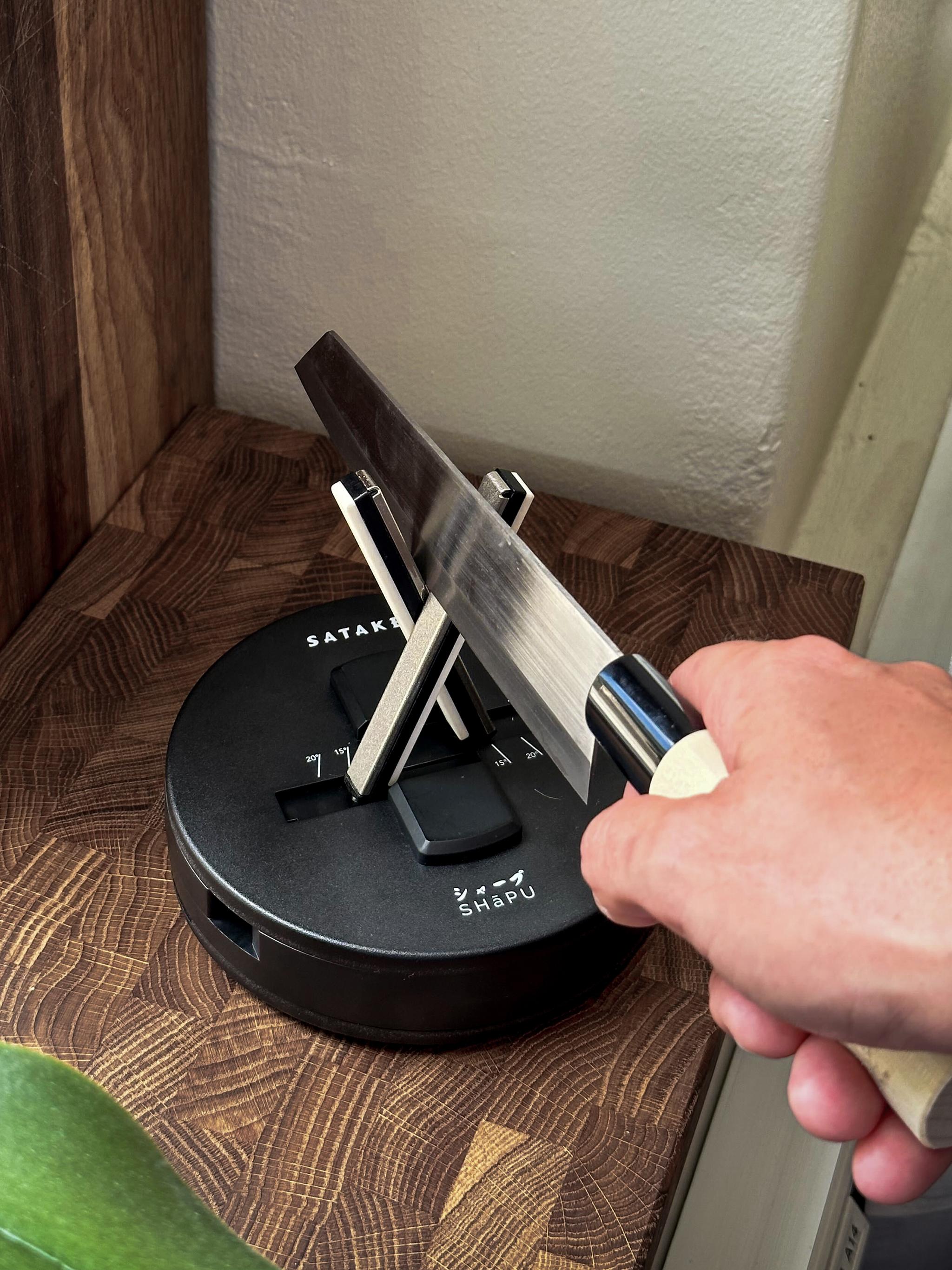 En persons hand styr en kniv in i en svart knivslipare som vilar på ett slaktblock av trä.