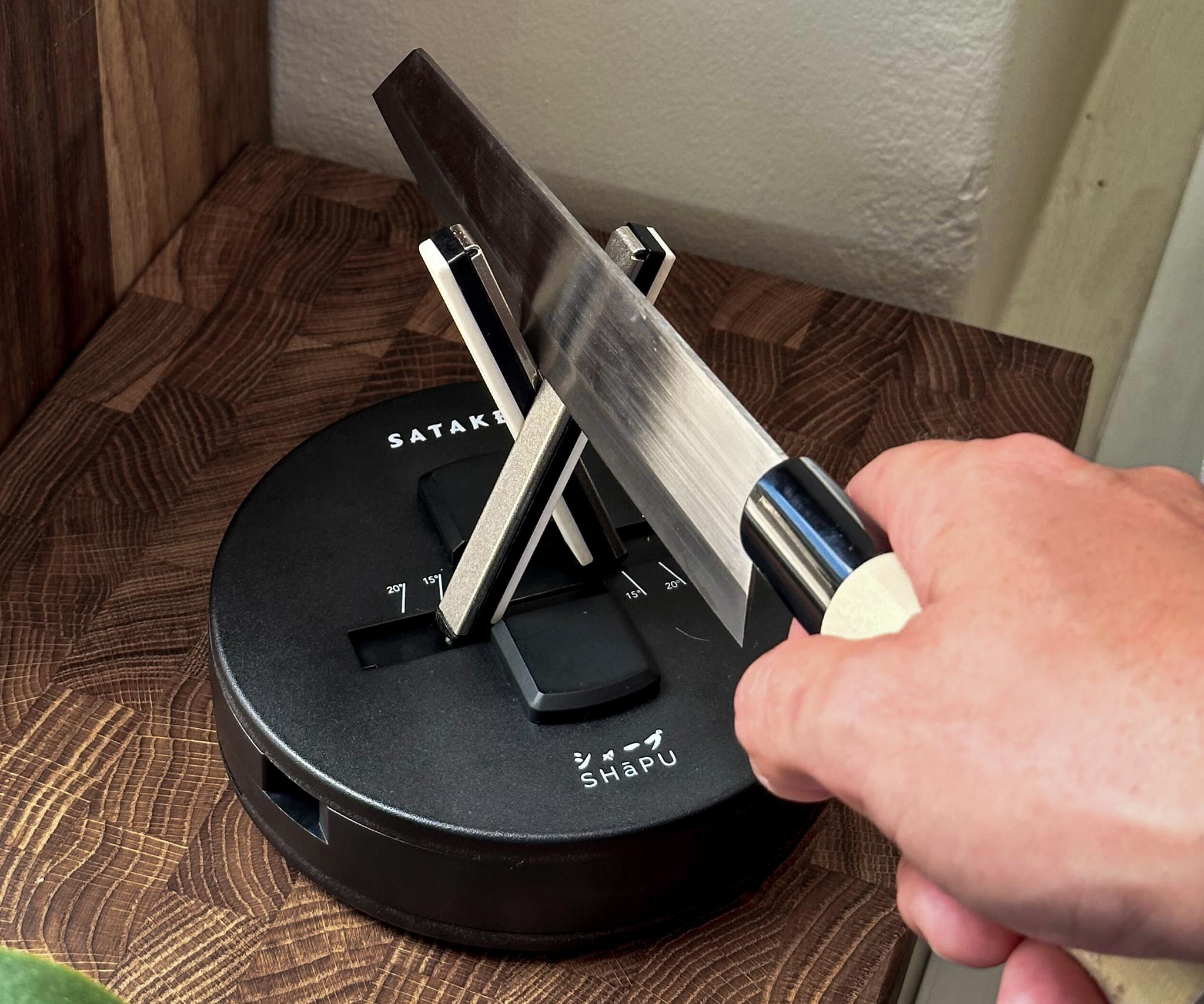 "En hand vässar en kniv på en svart Satake-knivslip som står på en träskärbräda