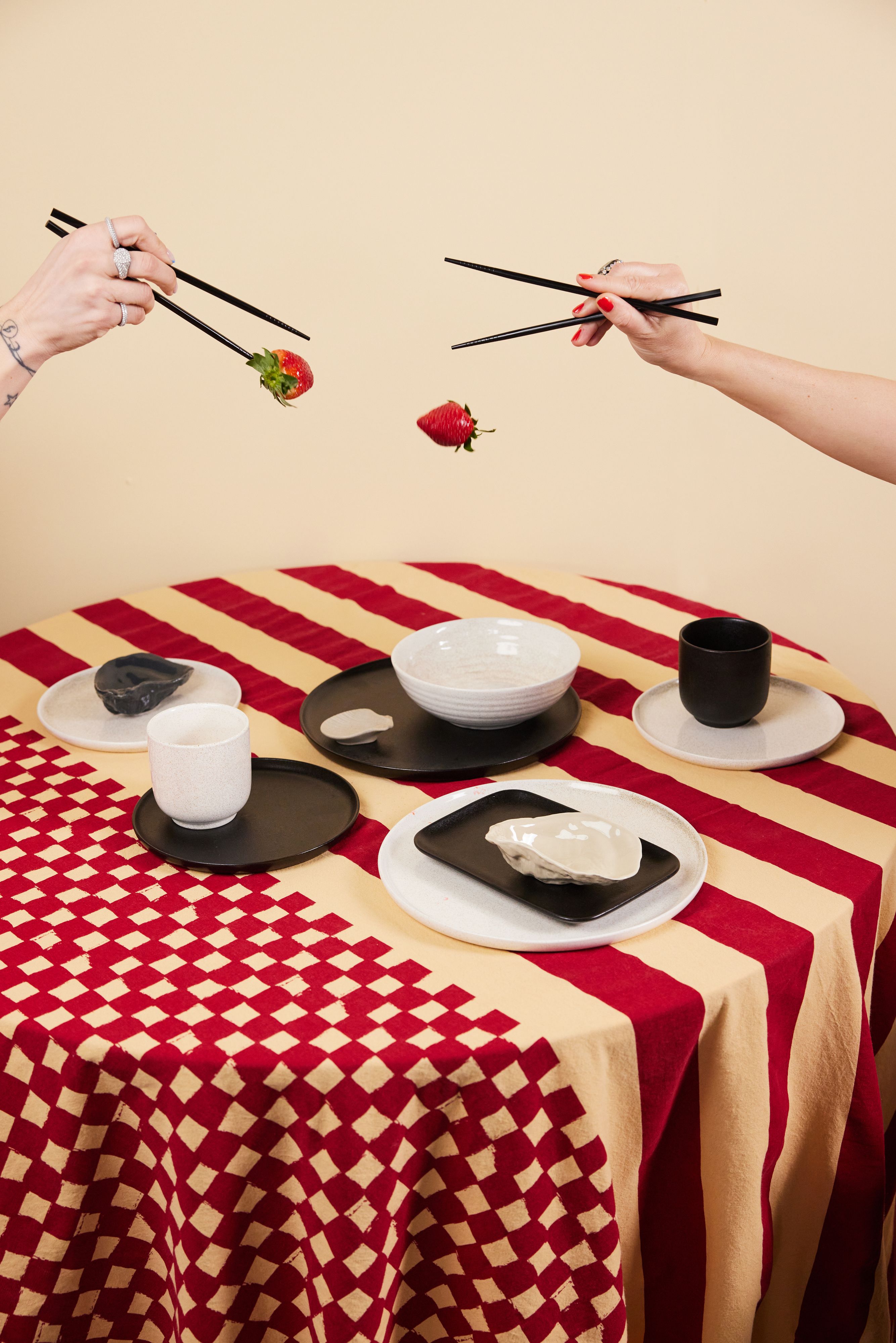 Två händer använder ätpinnar för att lekfullt släppa och hålla jordgubbar ovanför ett bord dukat med moderna tallrikar i svart, vitt och beige på en röd- och beigemönstrad duk