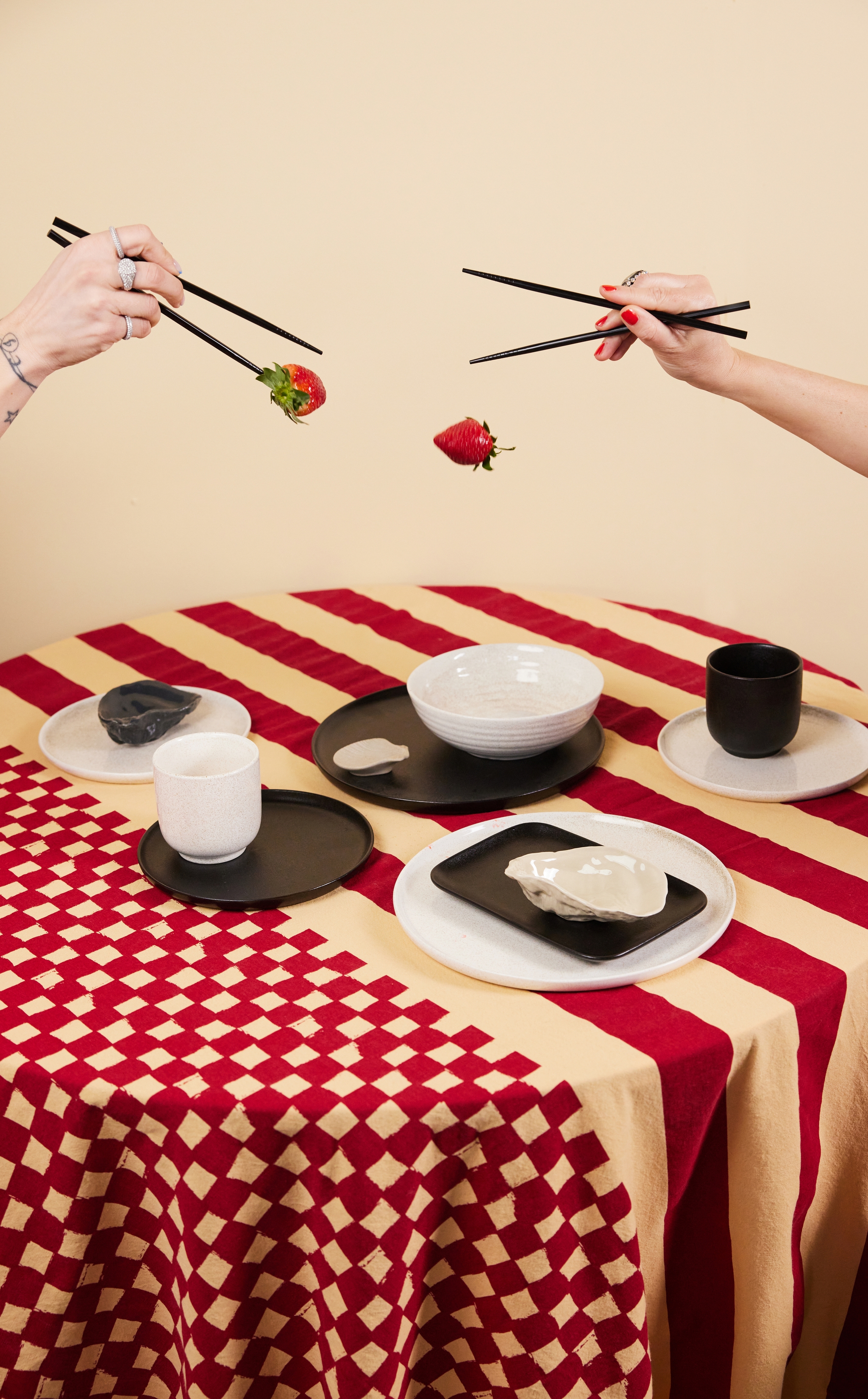 Två händer använder ätpinnar för att lekfullt släppa och hålla jordgubbar ovanför ett bord dukat med moderna tallrikar i svart, vitt och beige på en röd- och beigemönstrad duk