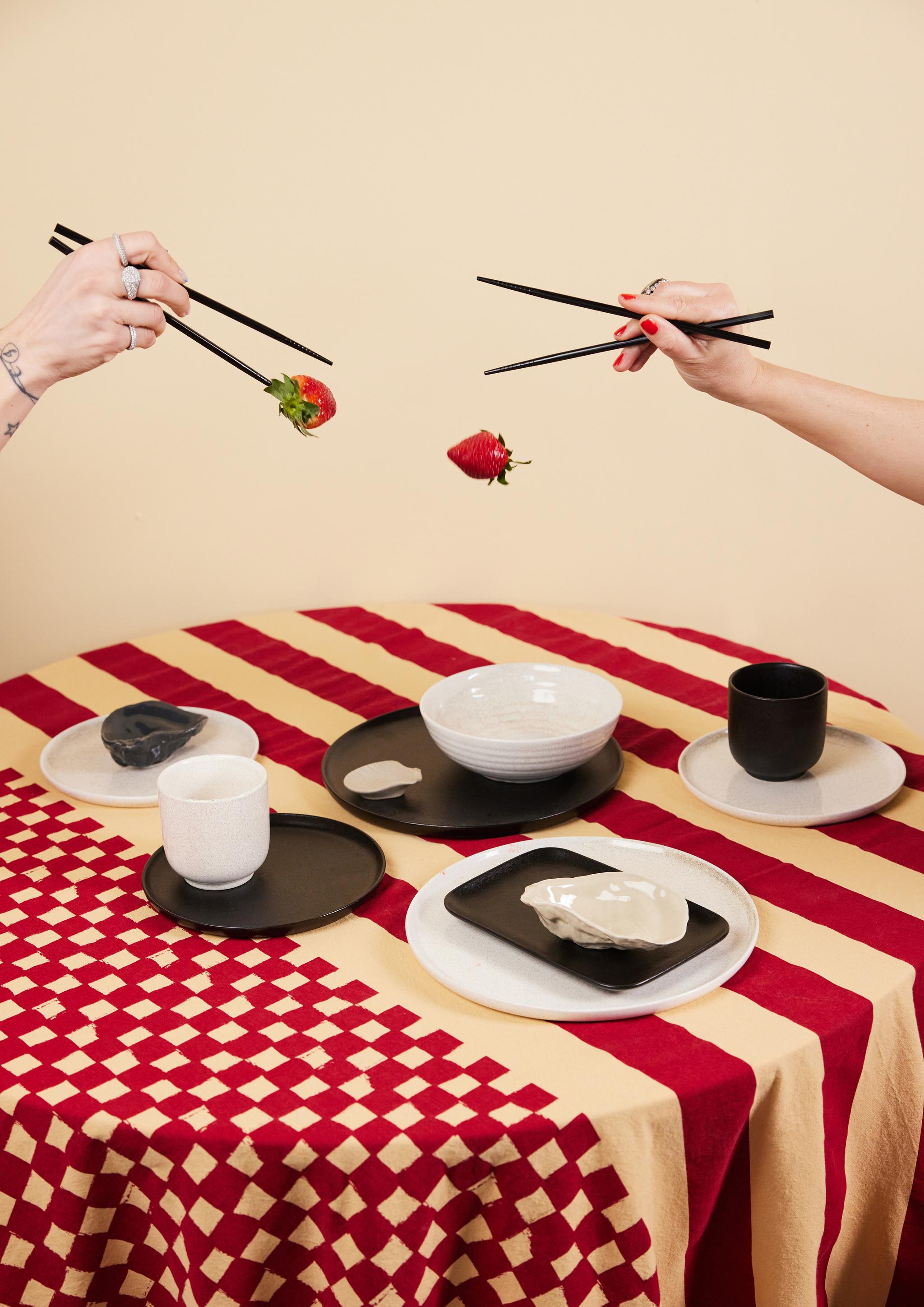 Två händer använder ätpinnar för att lekfullt släppa och hålla jordgubbar ovanför ett bord dukat med moderna tallrikar i svart, vitt och beige på en röd- och beigemönstrad duk