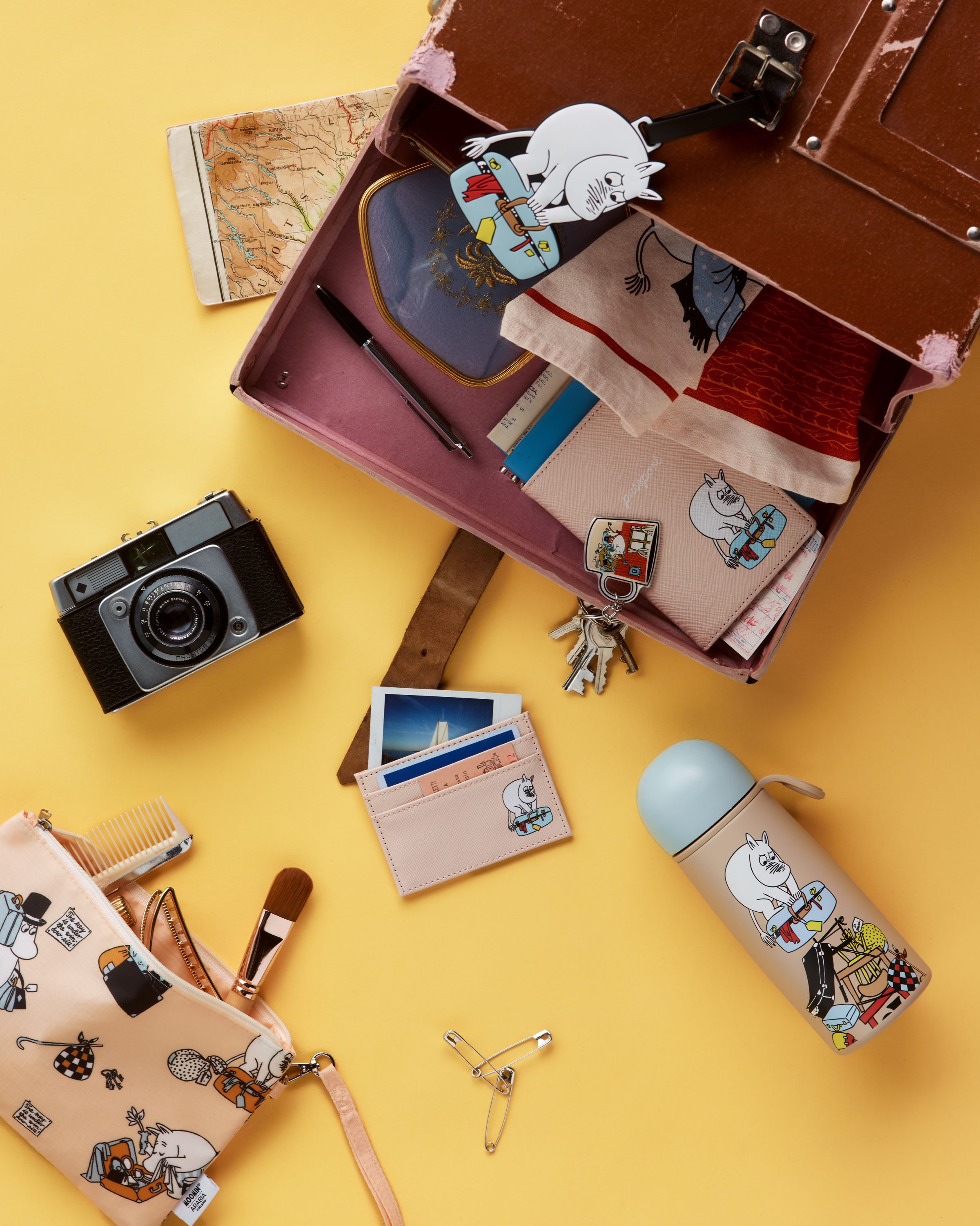 A vintage camera and an open pink container holding a passport, keys, and a pen, all on a yellow background.