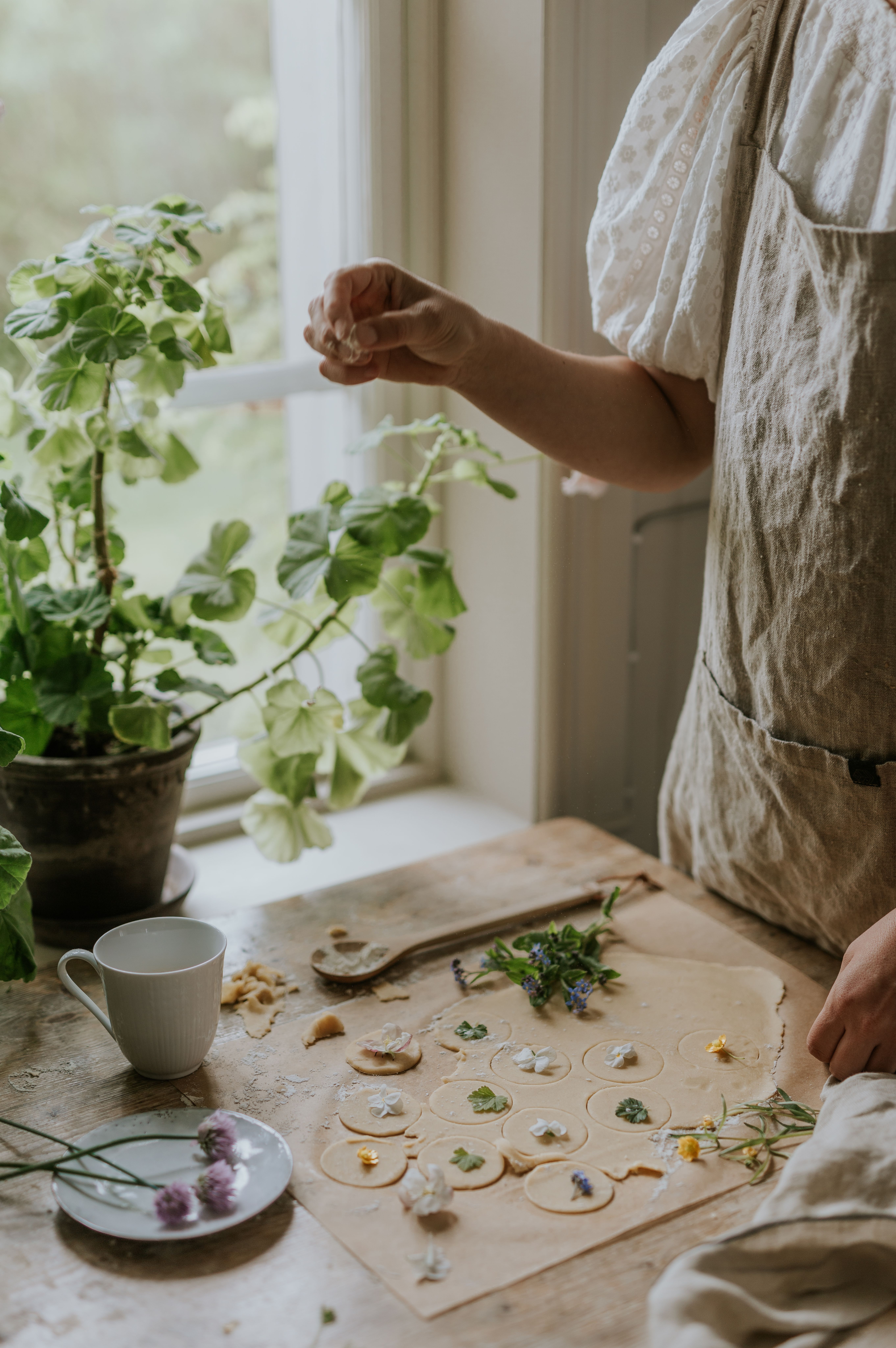 Person bakar småkakor med ätbara blommor på ett rustikt köksbord vid ett fönster med gröna krukväxter.