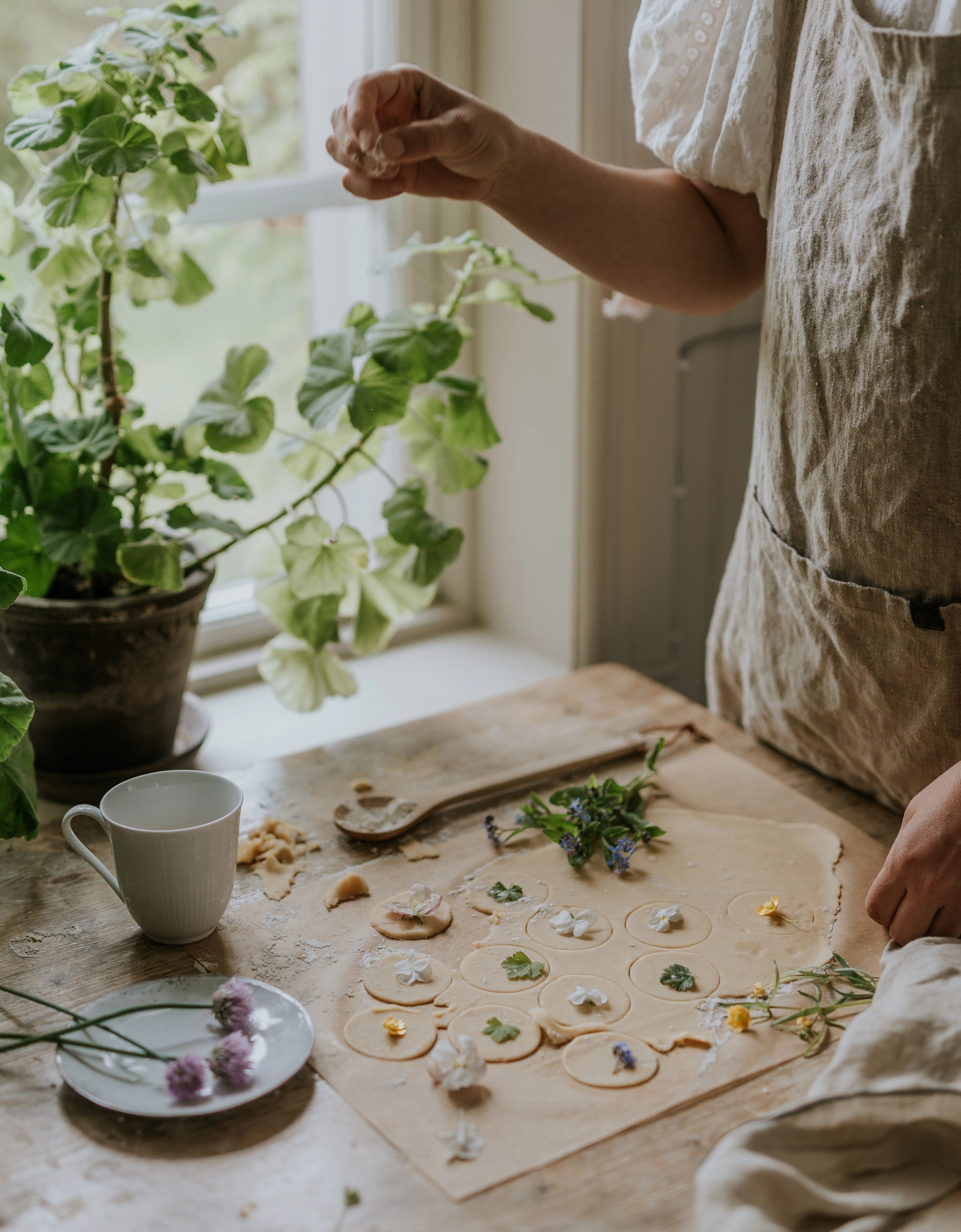 Person dekorerar hemmagjord deg med ätbara blommor vid ett ljust köksfönster med gröna krukväxter.