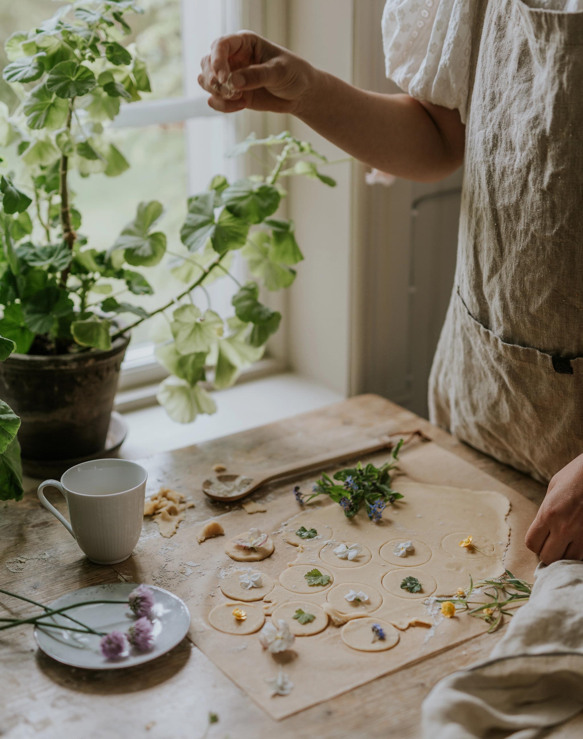 Person bakar småkakor med ätbara blommor på ett rustikt köksbord vid ett fönster med gröna krukväxter.