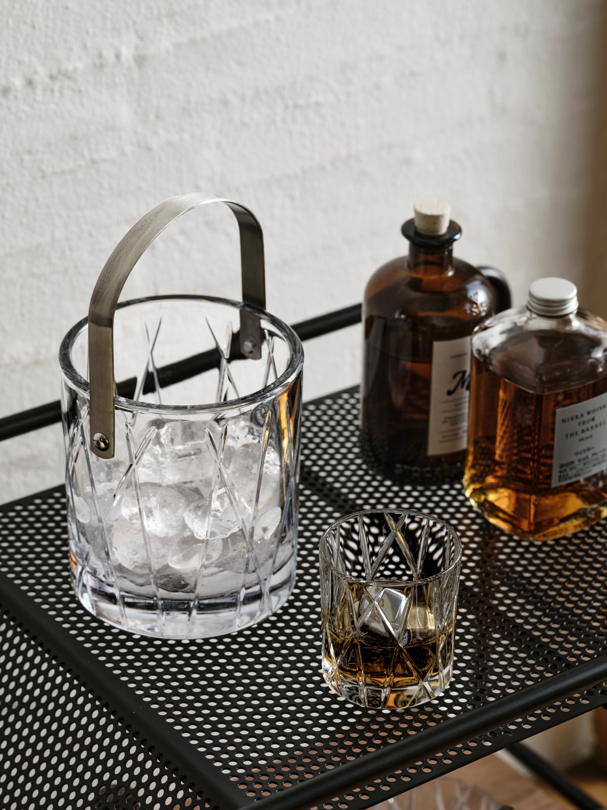 En kristallishink med is, ett whiskyglas och två flaskor på en svart perforerad barvagn.