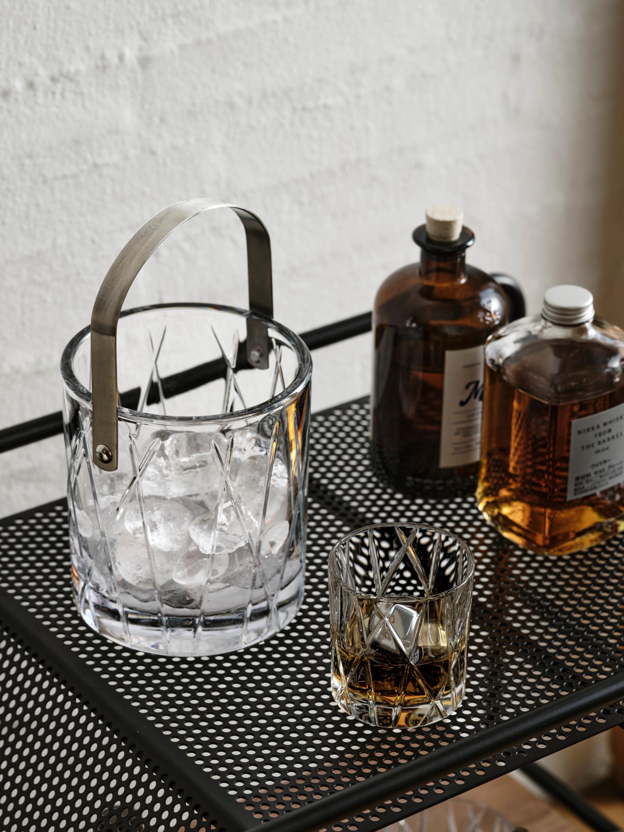 En kristallishink med is, ett whiskyglas och två flaskor på en svart perforerad barvagn.