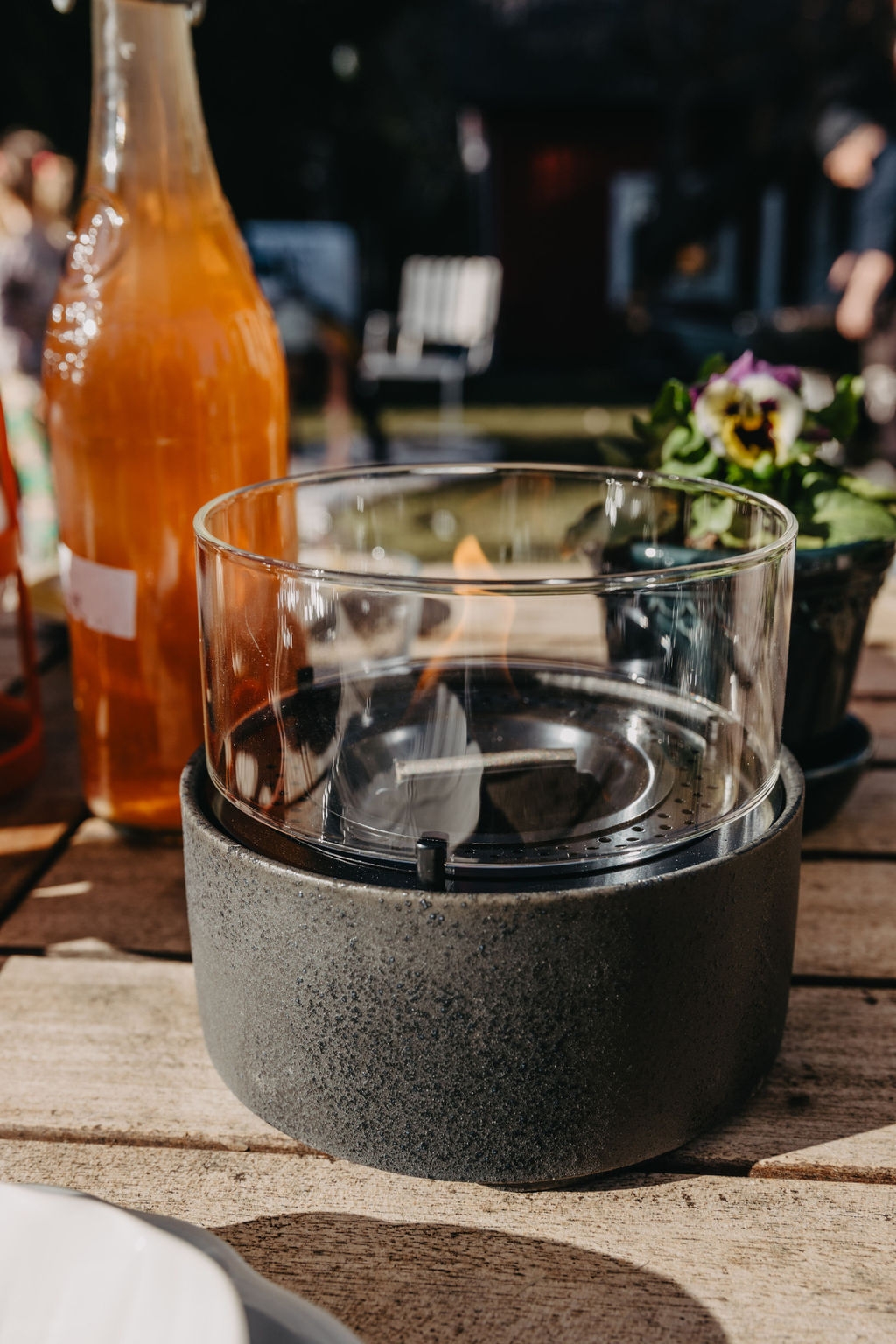 En modern bordseld i glas och betong står på ett solbelyst trädäck bredvid en flaska dryck och en blomkruka.