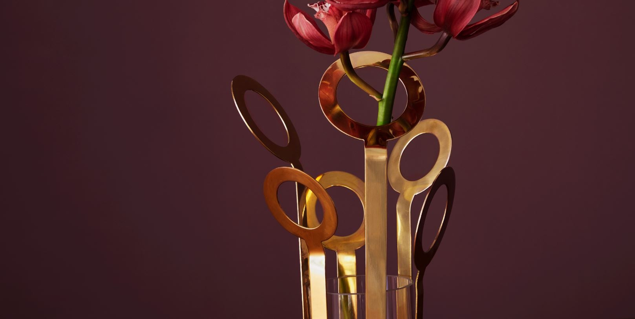 Vasen Fanny i mässing med ringformade detaljer och röda blommor mot mörkt vinrött bakgrund