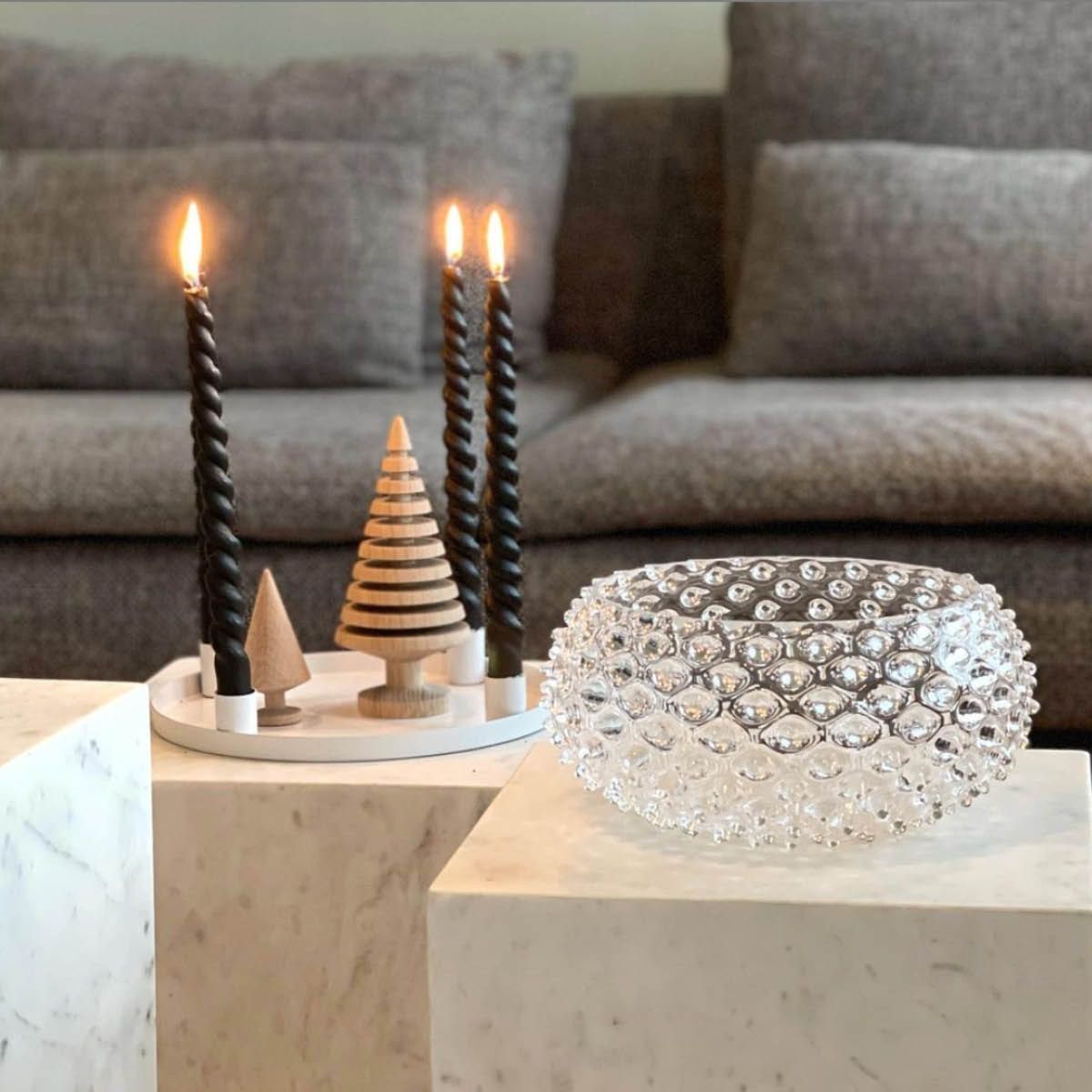 Svarta spiralformade ljus, dekorativa trägranar och en skål i bubbligt glas på marmorbord framför en grå soffa.