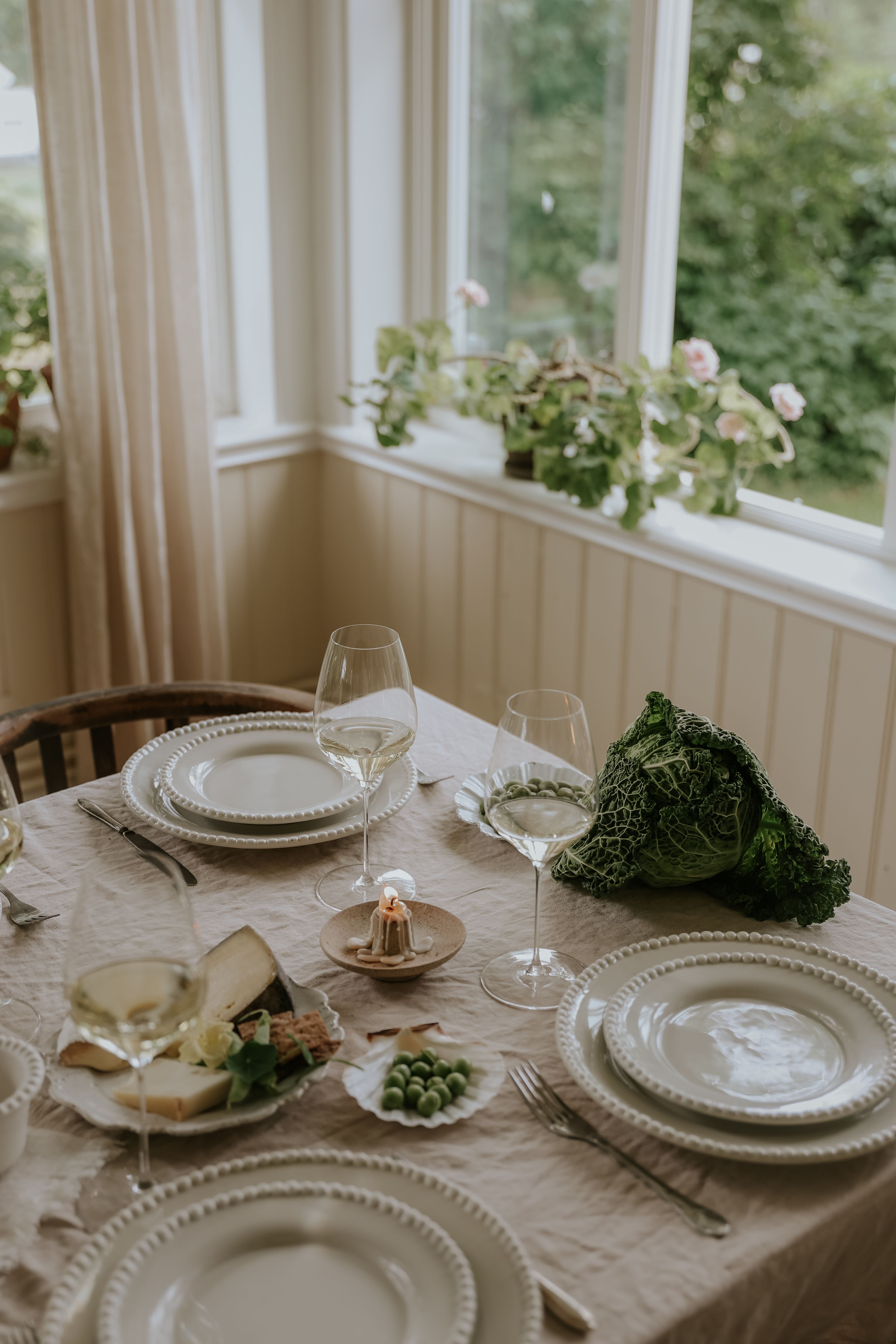 Ett matbord dukat med vita pärltallrikar, vinglas, ett tänt ljus, ost, ärtor och ett kålhuvud, med ett fönster och växter i bakgrunden.