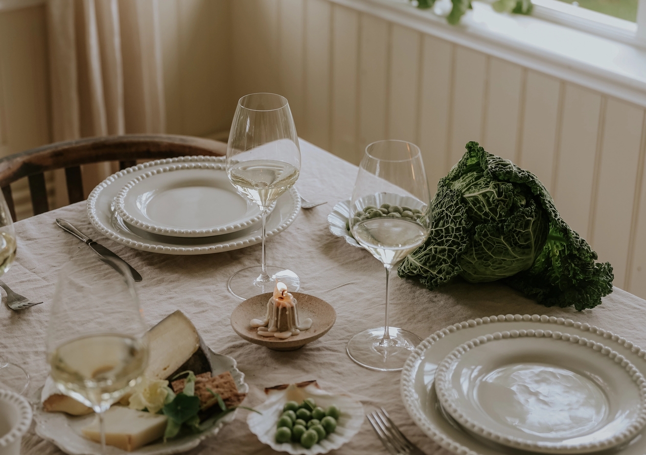 Vackert dukat matbord i lantlig stil med vin, ost, grönkål och utsikt mot grönskande trädgård genom stora fönster.