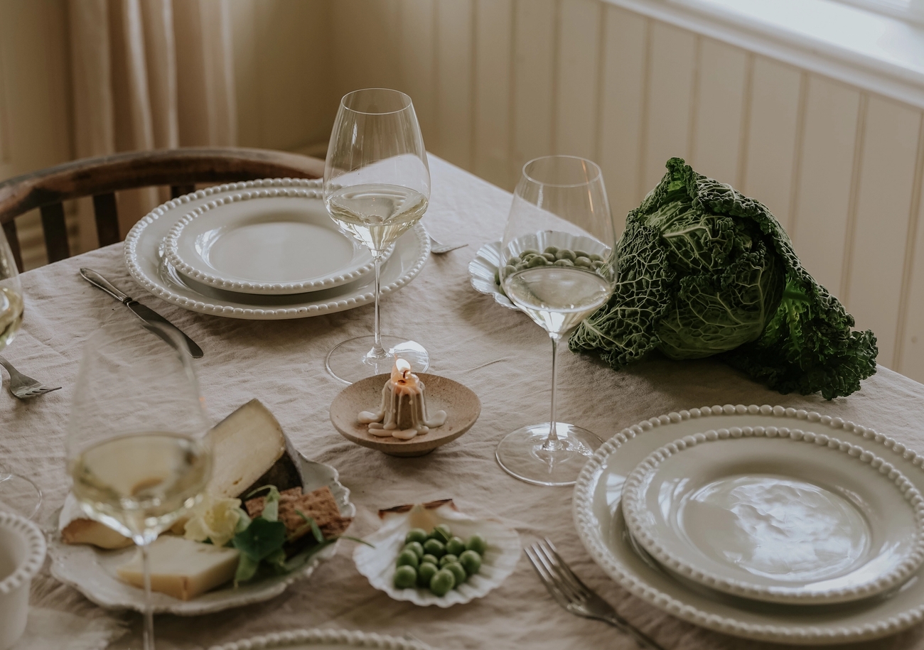 Ljus och lantlig dukning med vin, ost och grönkål på ett bord vid ett fönster med gröna växter.