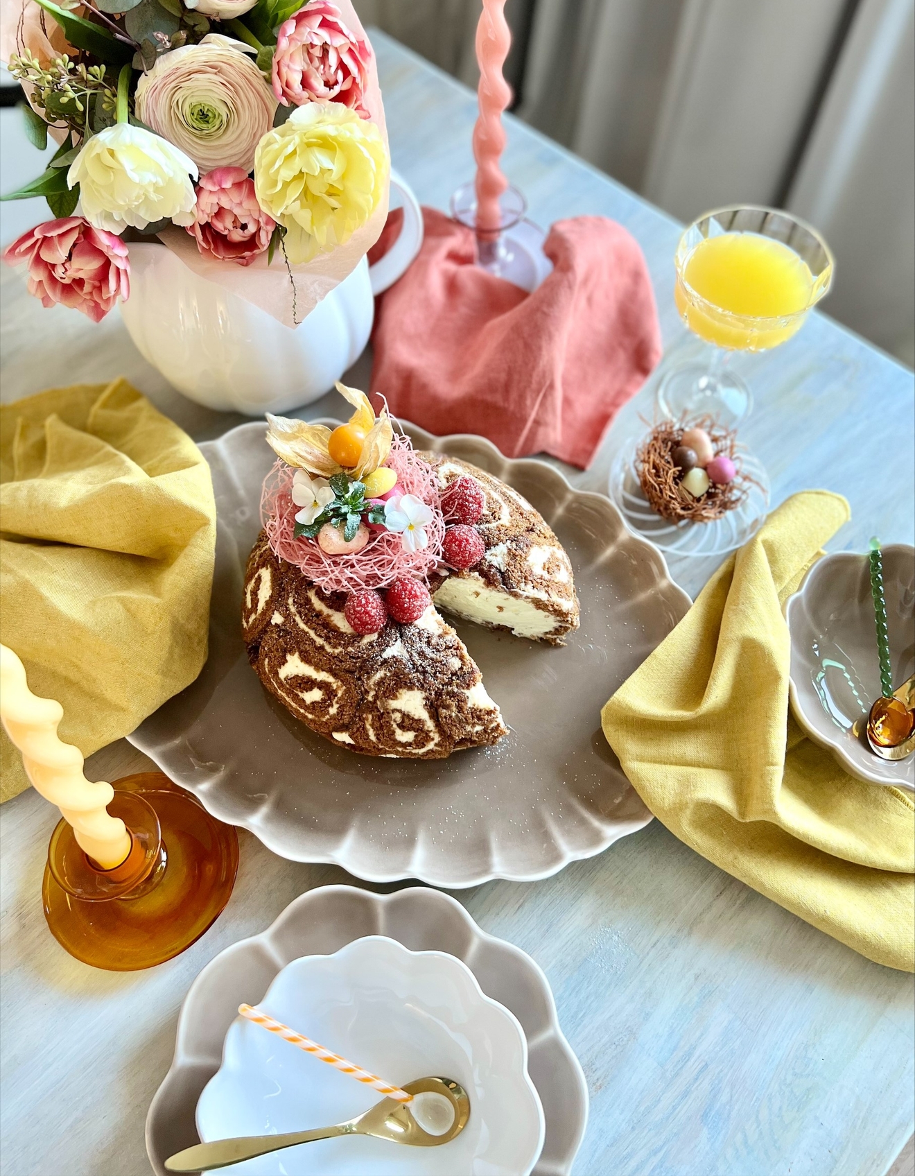 Ett festligt bord visar en skivad chokladrulltårta med hallon, en bukett rosa och gula blommor, vågiga ljus, apelsinjuice och ett litet bo med chokladägg