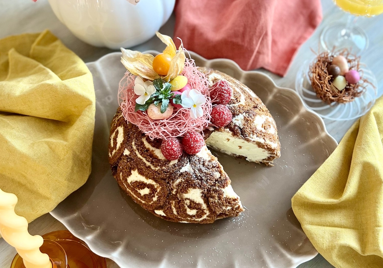 Ett festligt bord visar en skivad chokladrulltårta med hallon, en bukett rosa och gula blommor, vågiga ljus, apelsinjuice och ett litet bo med chokladägg