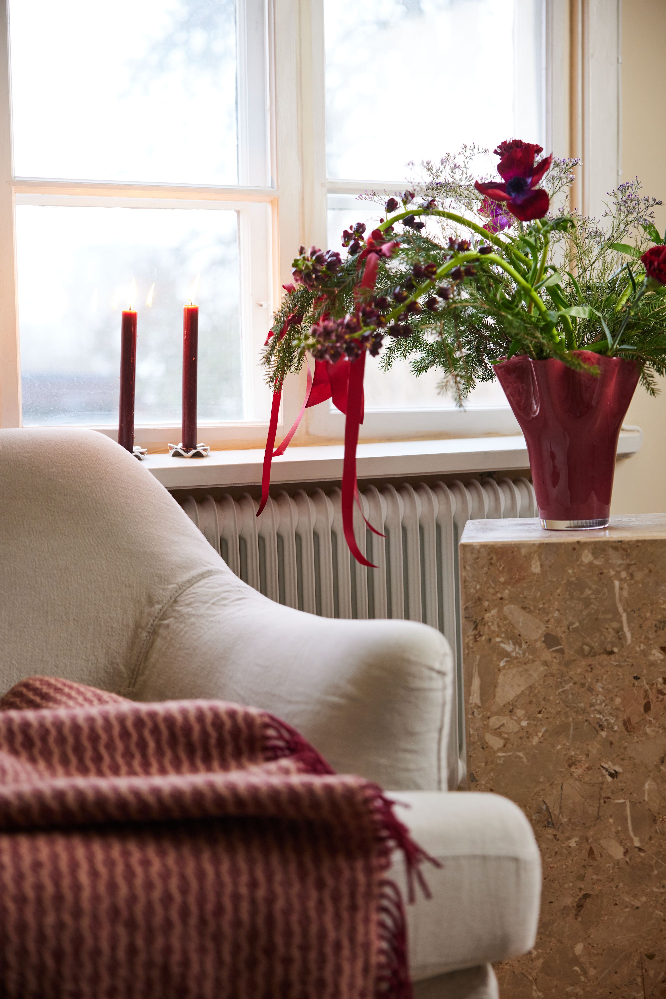 Mysig vardagsrumshörna med vit fåtölj, röd pläd, tända ljus och julig blomsterbukett i röd vas vid fönstret.