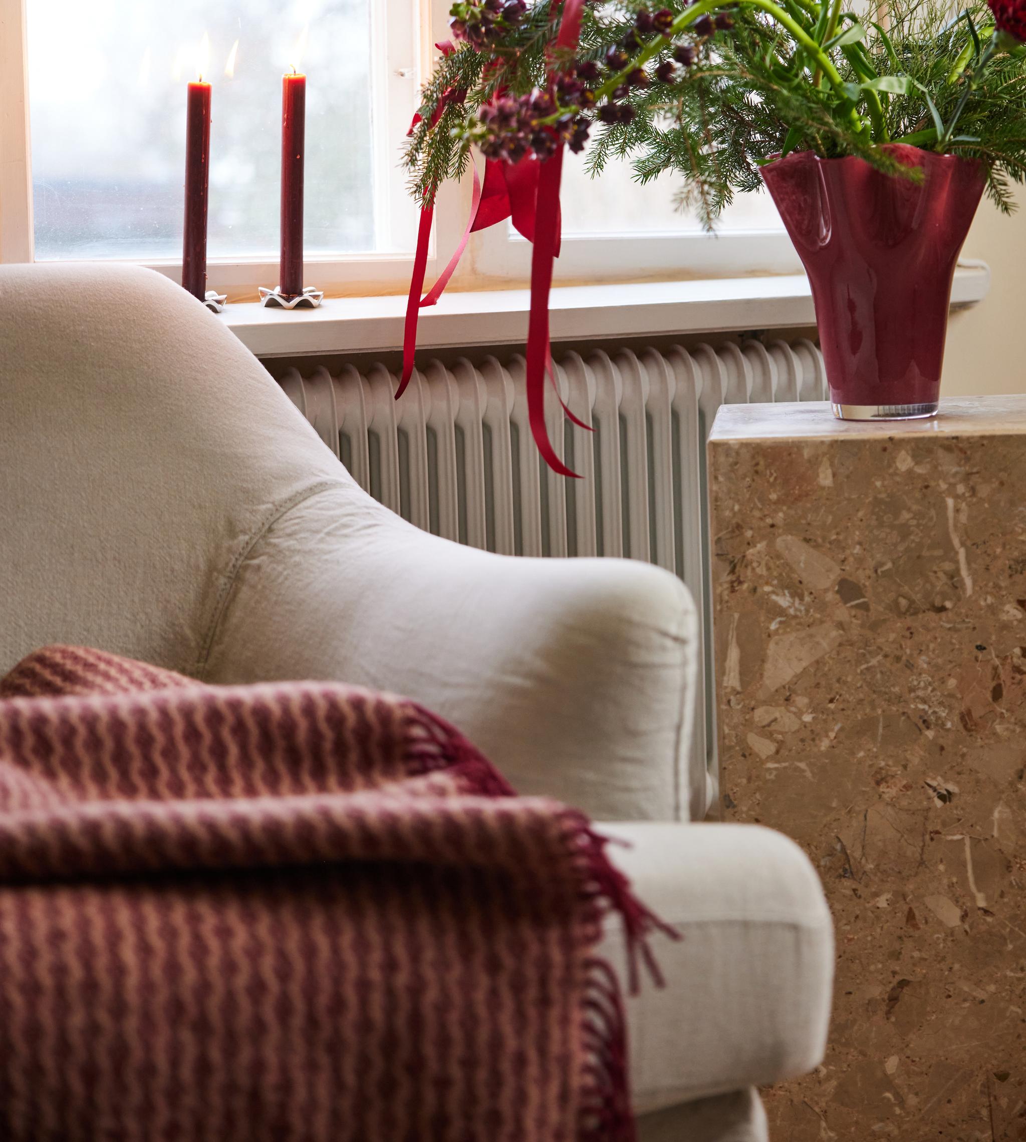 Mysig vardagsrumshörna med vit fåtölj, röd pläd, tända ljus och julig blomsterbukett i röd vas vid fönstret.