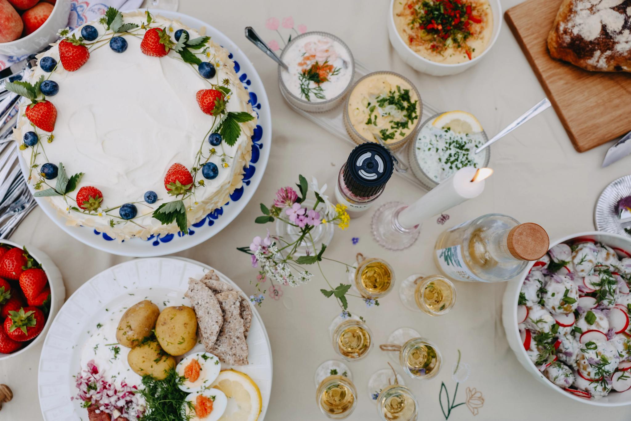 Somrig svensk festdukning med jordgubbstårta, sill, färskpotatis, snaps och blommor på bordet.