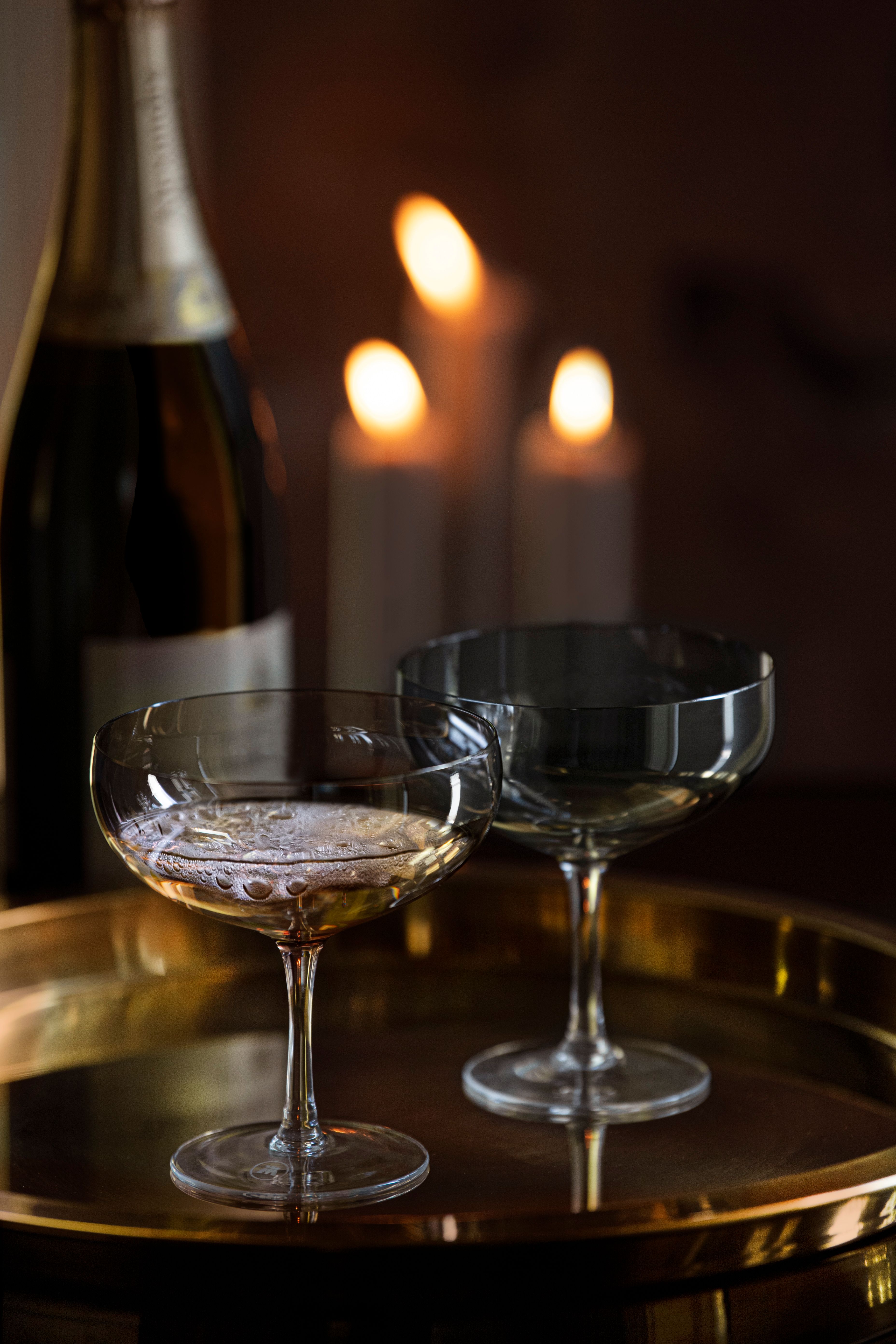 Två champagneglas och en flaska på en guldfärgad bricka framför tre tända ljus.