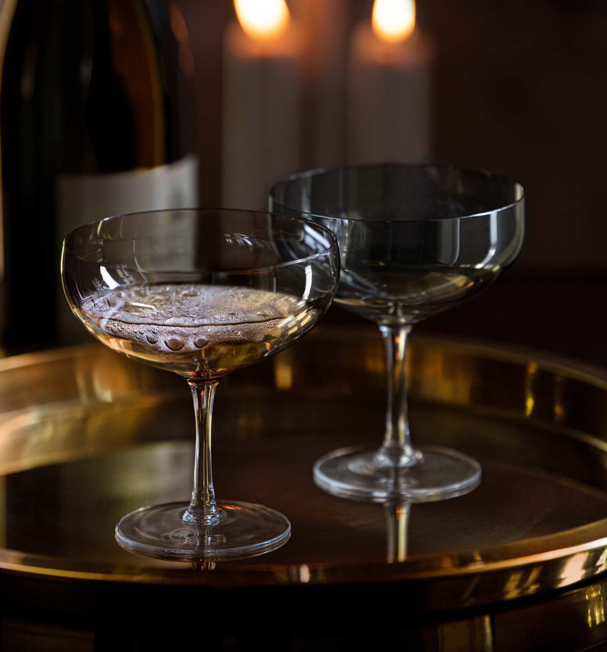 Två champagneglas och en flaska på en guldfärgad bricka framför tre tända ljus.