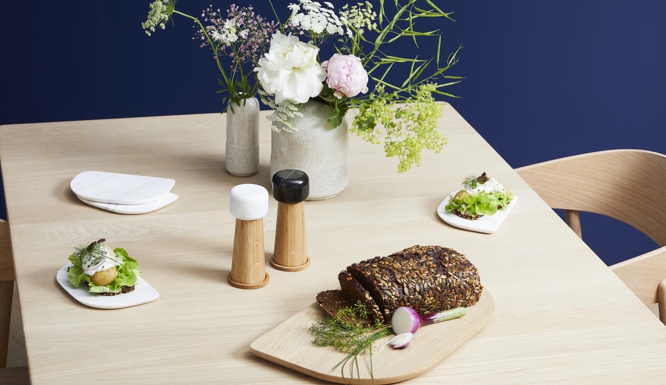 Ett modernt träbord är dukat med blommor, salt- och pepparkvarnar, skivat bröd med frön och små aptitretare, mot en mörkblå vägg.