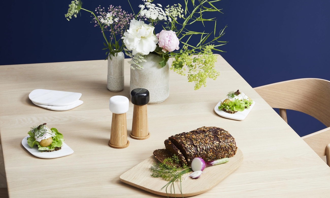A wooden table set with a vase of flowers, two minimalist salt and pepper grinders, sliced seeded bread, and two open-faced sandwiches.