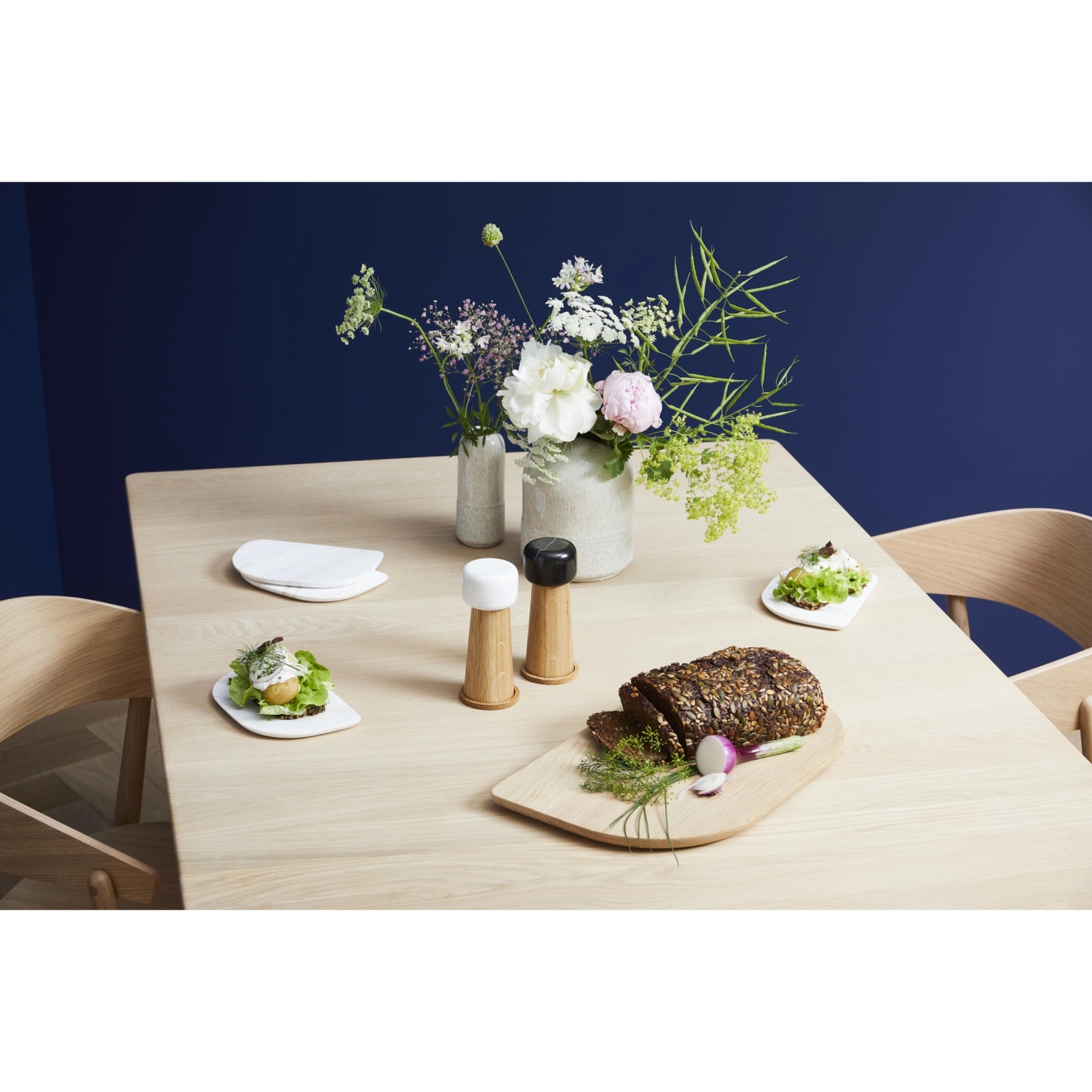 Et moderne trebord er dekket med blomster, salt- og pepperkverner, oppskåret brød med frø og små forretter, mot en mørkeblå vegg.