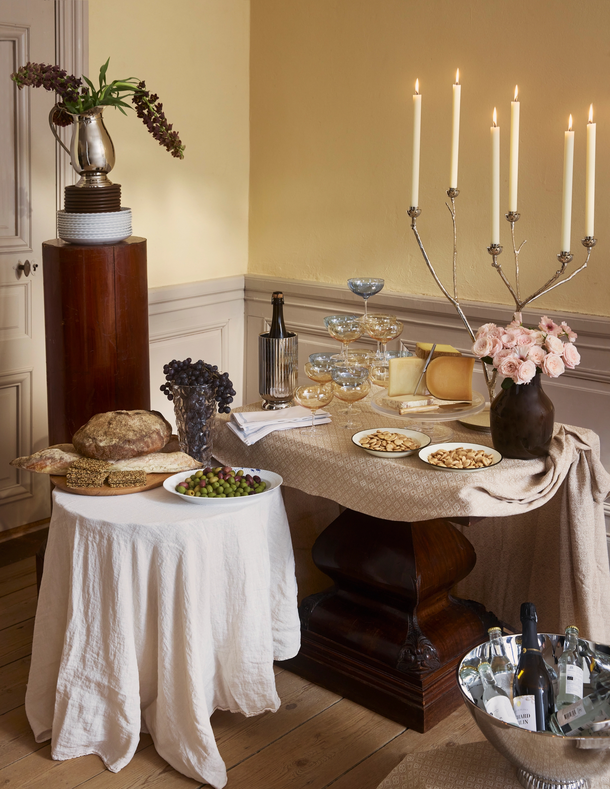 En elegant buffé med ost, bröd, oliver, drycker, tända ljus och blomsterarrangemang på två bord