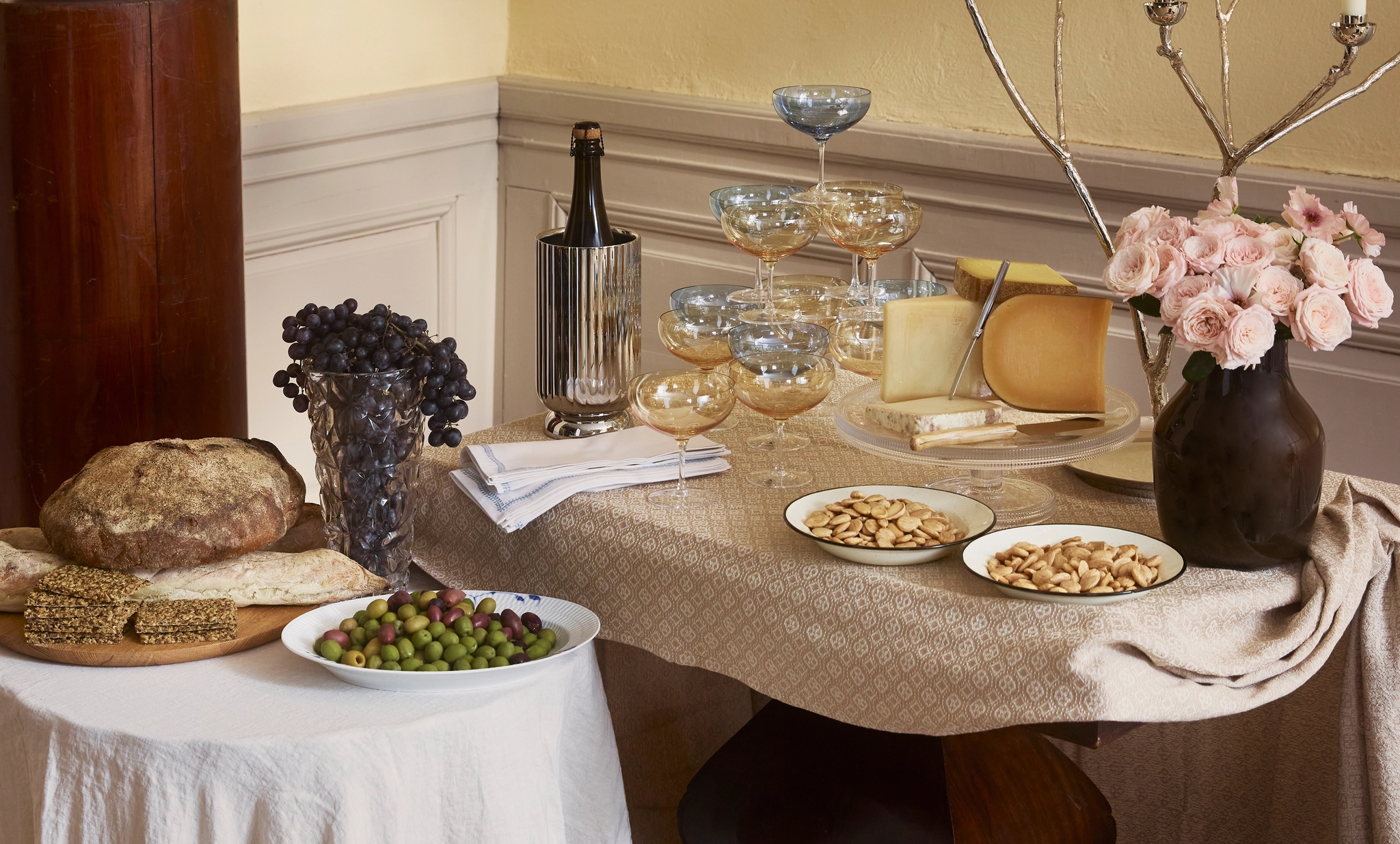 En elegant buffé med ost, bröd, oliver, drycker, tända ljus och blomsterarrangemang på två bord.