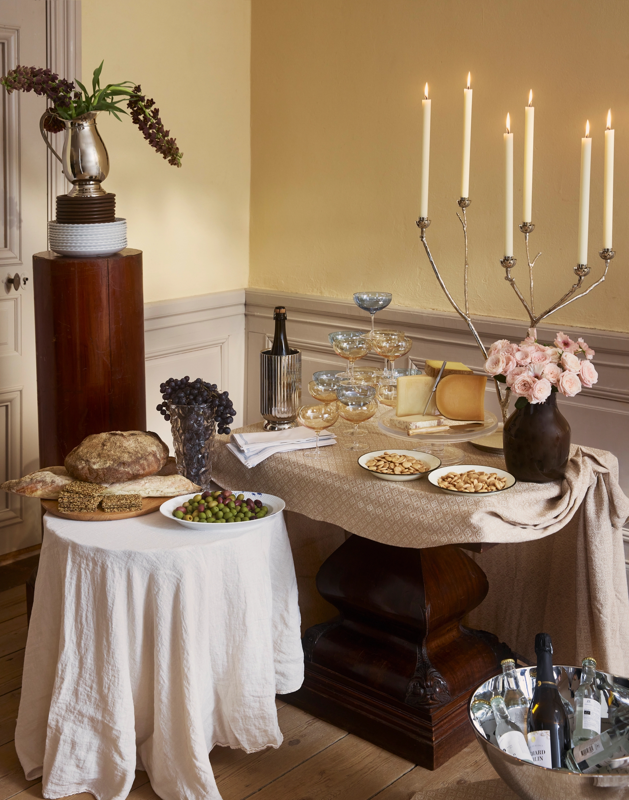 En elegant buffé med ost, bröd, oliver, drycker, tända ljus och blomsterarrangemang på två bord