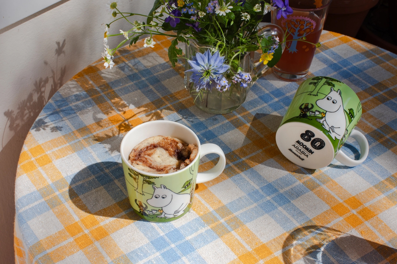 Two Moomin mugs on a yellow and blue plaid tablecloth, one holding a dessert, with a vase of wildflowers and a drink.