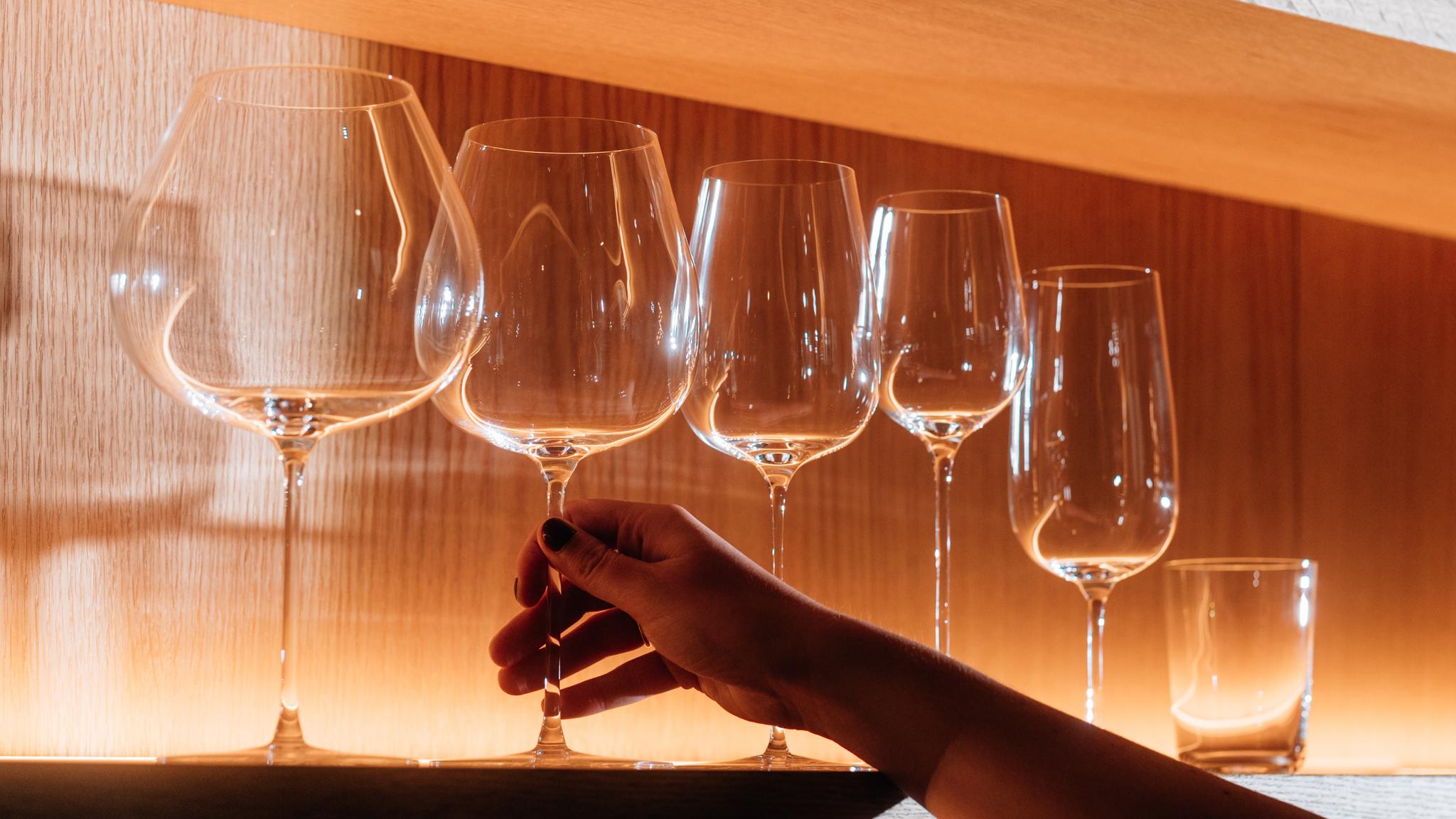 En hand sträcker sig efter ett klart vinglas bland flera andra på en varm, upplyst hylla.