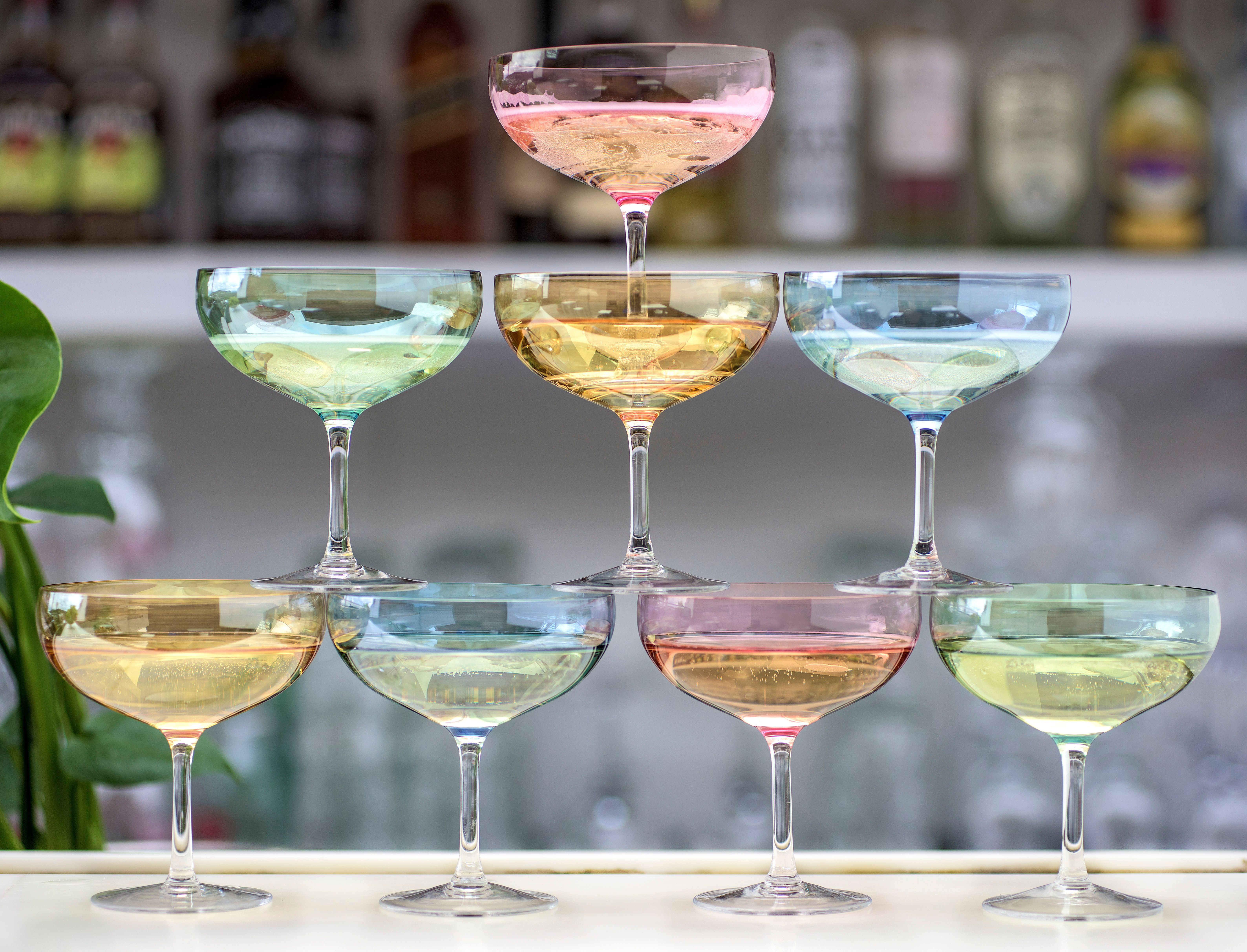 Fargerike champagneglass stablet i en pyramide på en bar med uskarpe flasker i bakgrunnen.