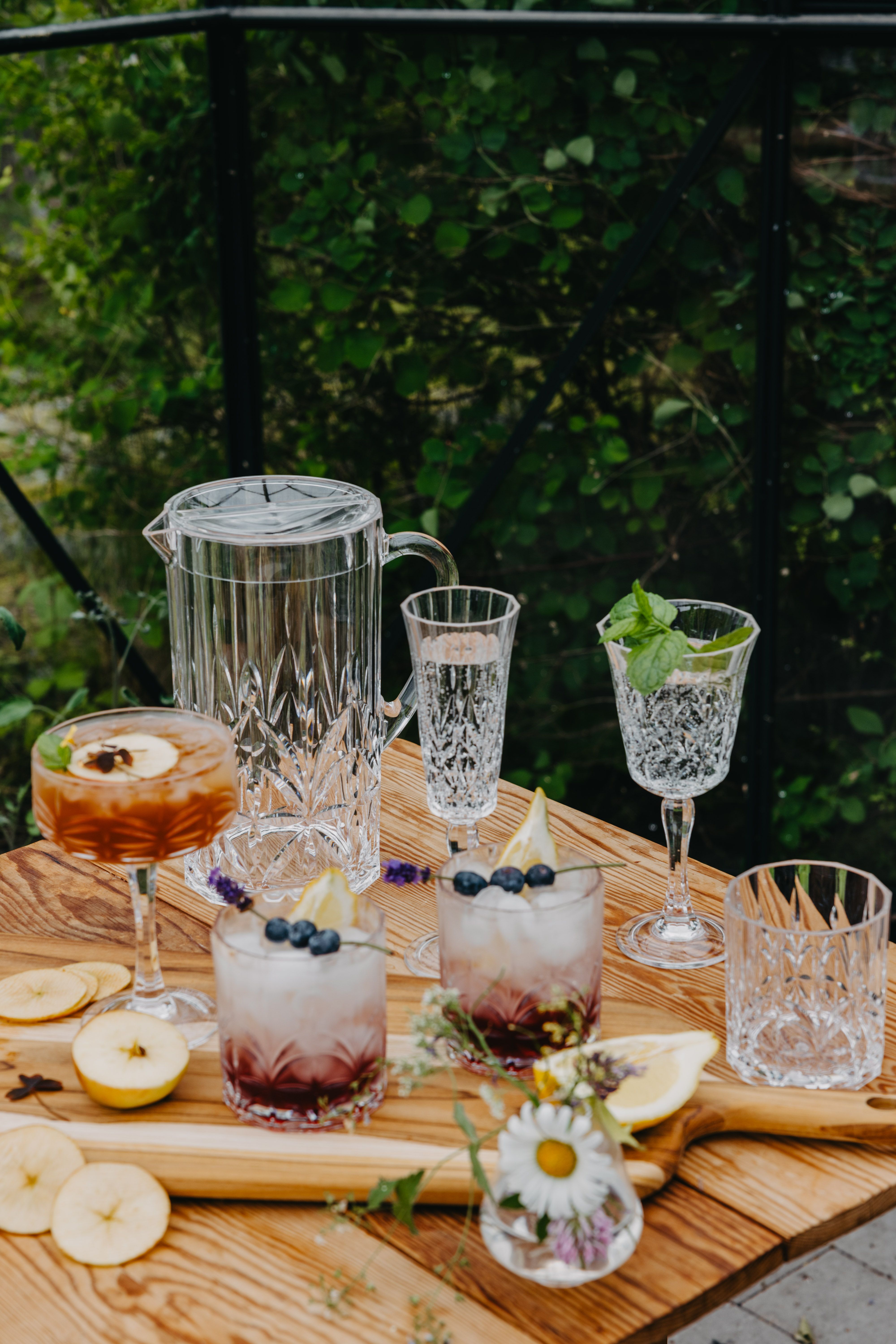 Somrig dukning utomhus med kristallglas och färgglada drinkar med frukt och bär på ett träbord.