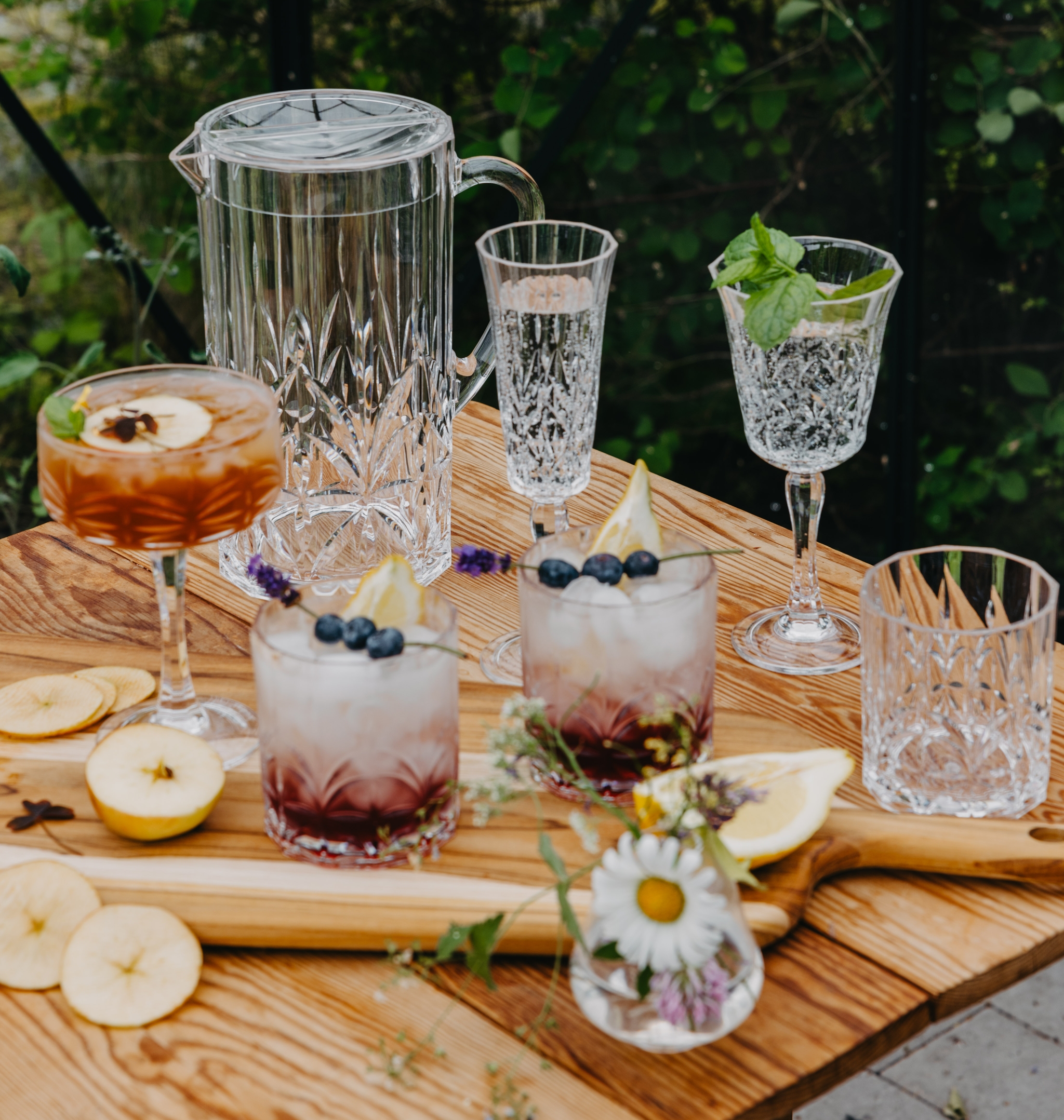 Somrig dukning utomhus med kristallglas och färgglada drinkar med bär, frukt och örter på ett rustikt träbord.