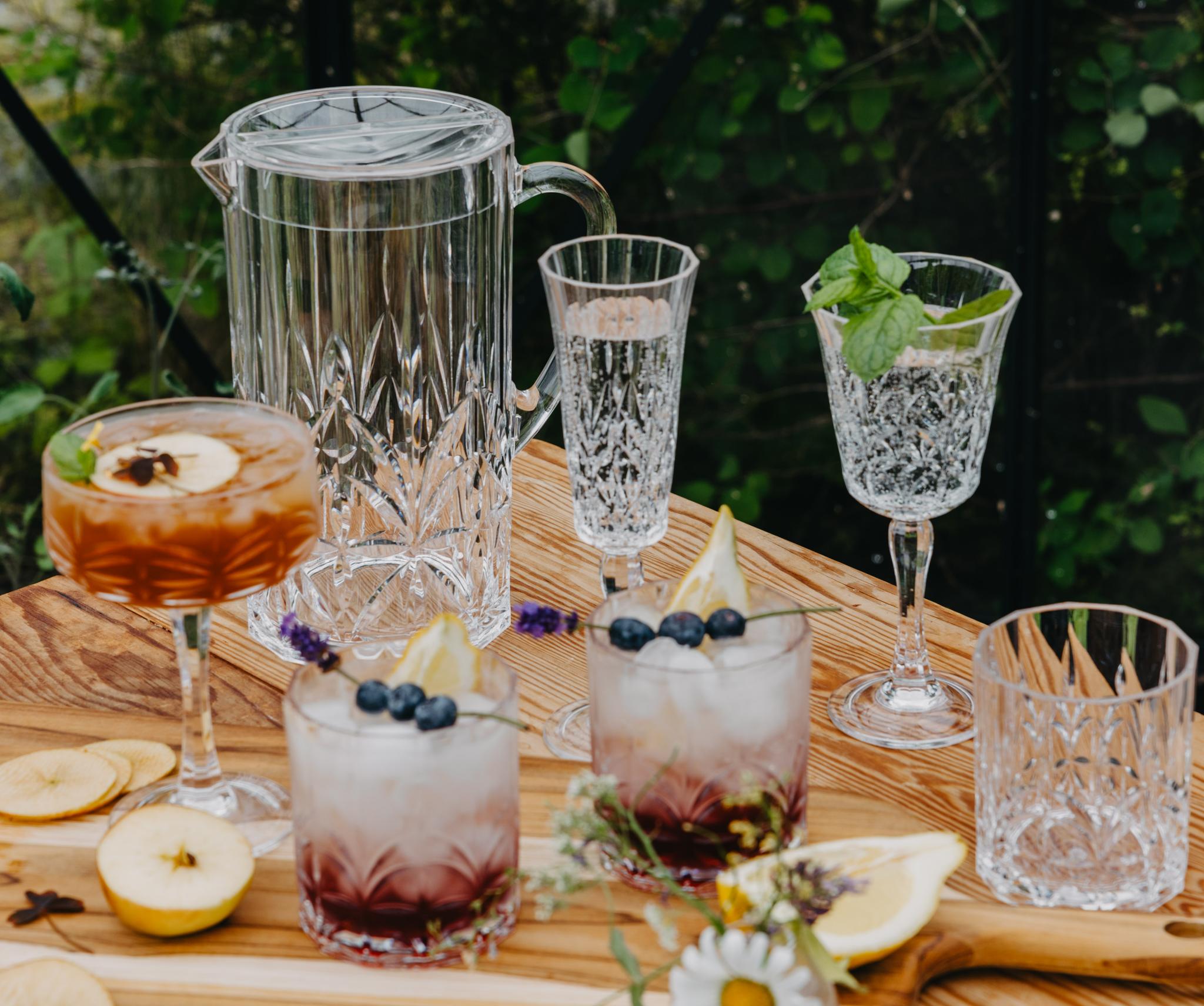 Sommerlig borddekking utendørs med krystallglass, friske drinker, frukt og blomster på et trebord.