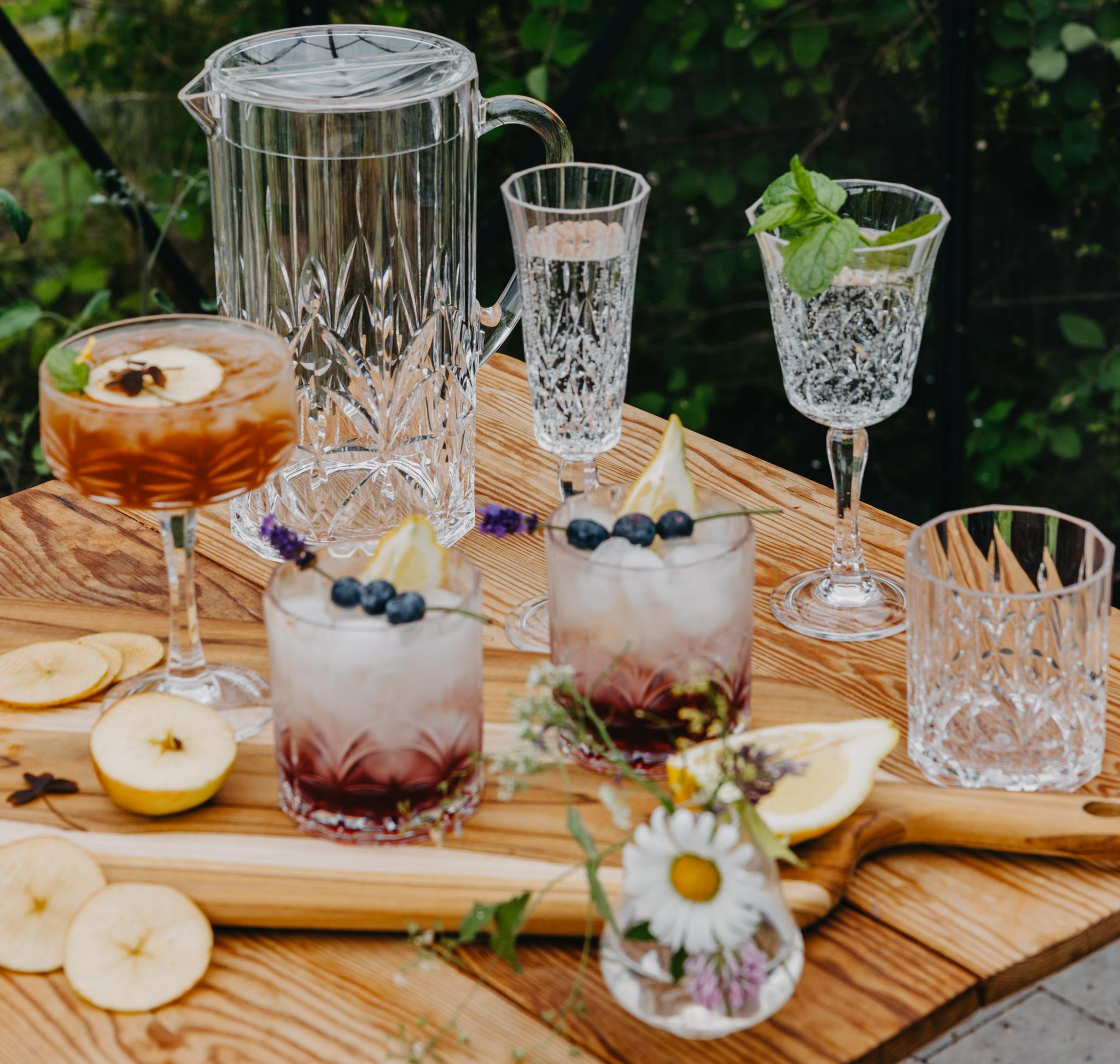 Somrig dukning utomhus med kristallglas och färgglada drinkar med frukt och bär på ett träbord.