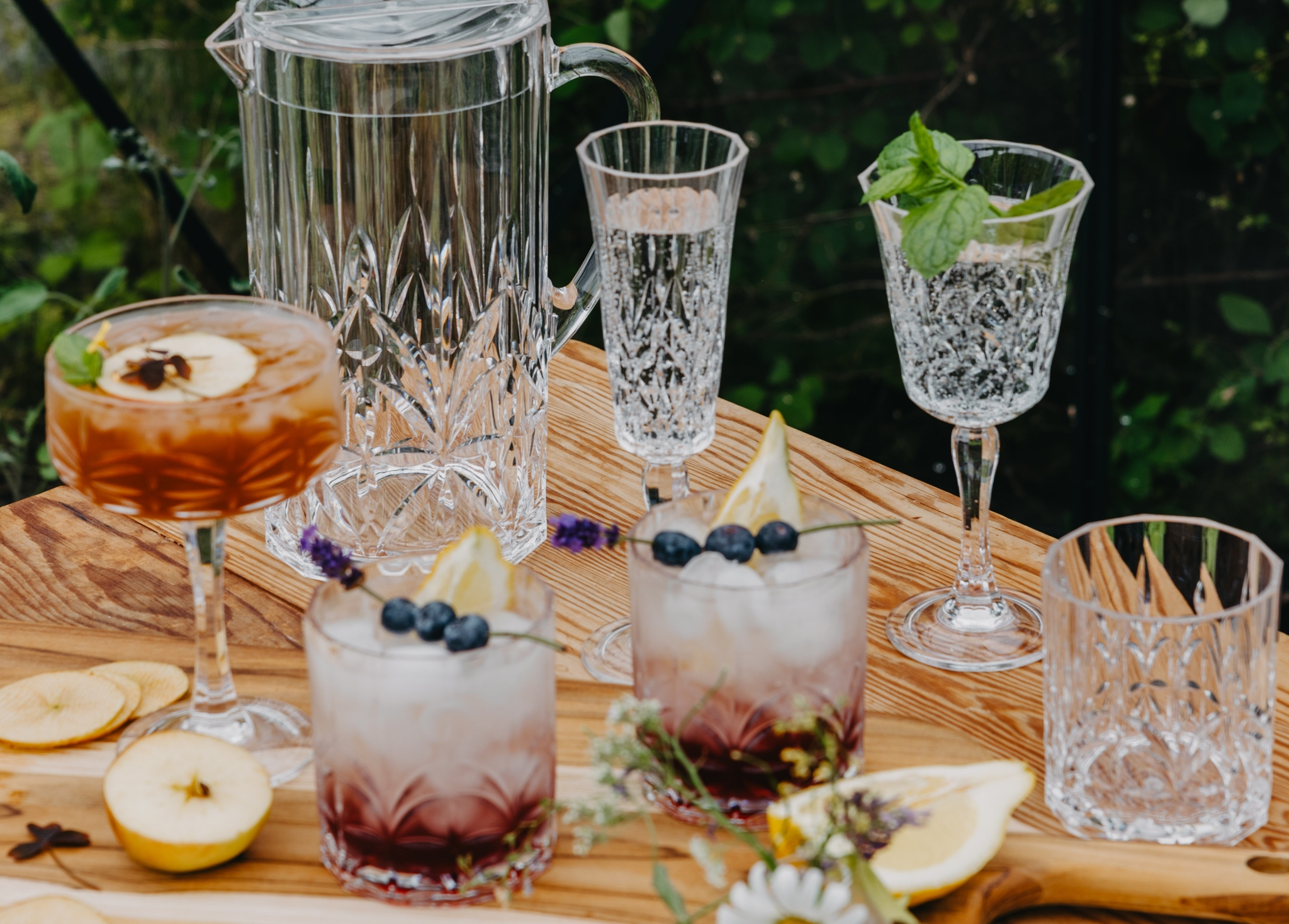 Sommerlig borddekking utendørs med krystallglass og friske cocktails pyntet med bær, urter og sitron på et rustikt trebord.