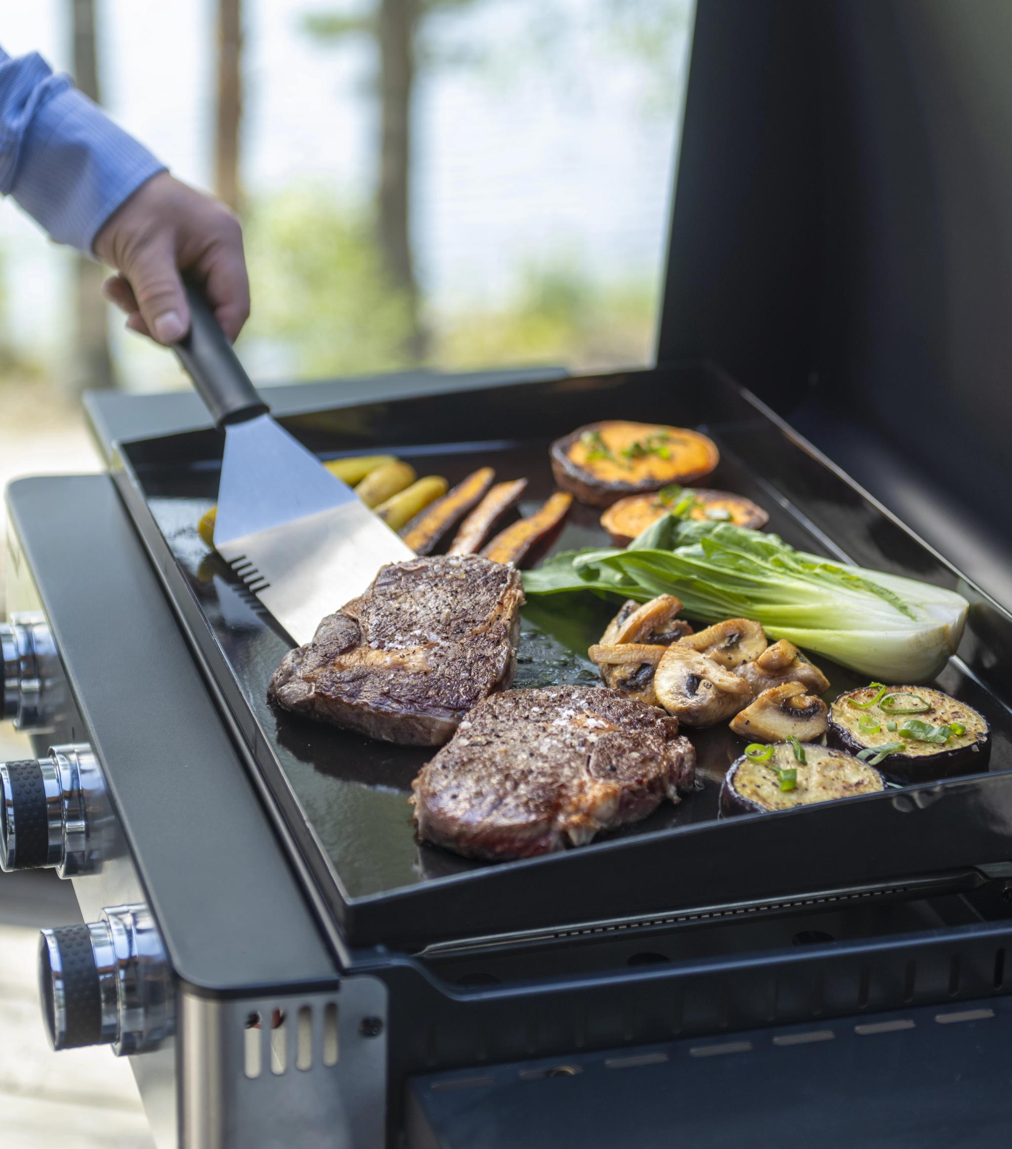 A hand flips steaks and vegetables on a griddle.