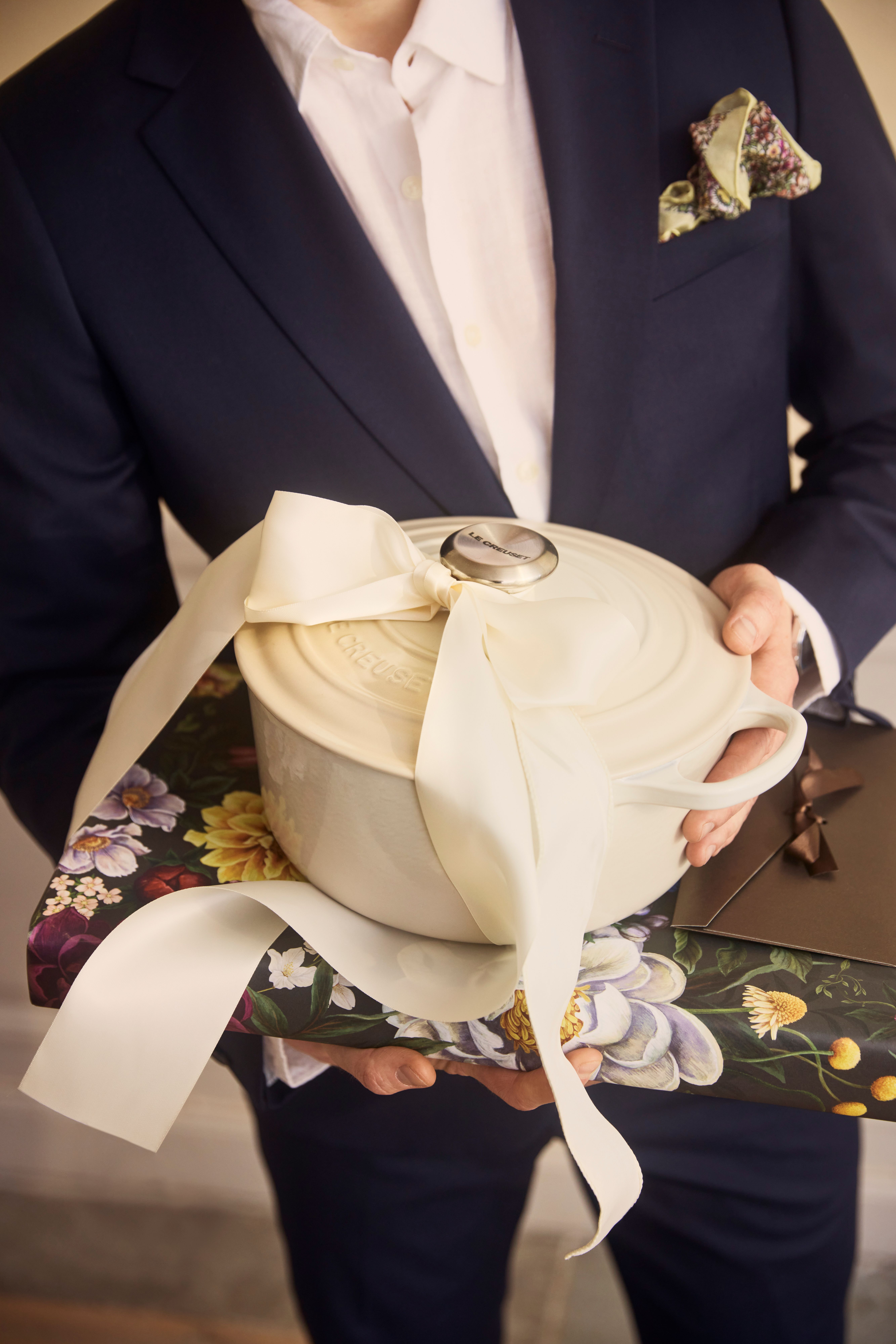 En man i kostym håller en vit Le Creuset-gryta med ett sidenband knutet i knoppen