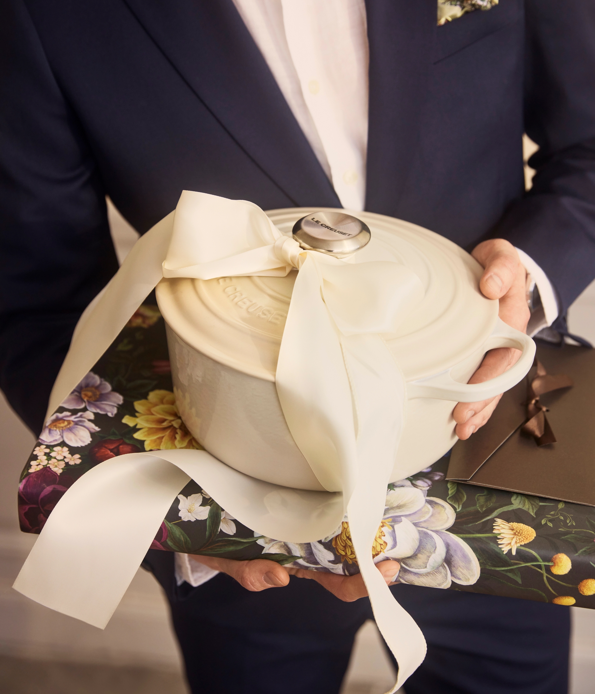 En person i dress holder en kremfarget Le Creuset-gryte med stor sløyfe oppå en blomstrete gaveeske.