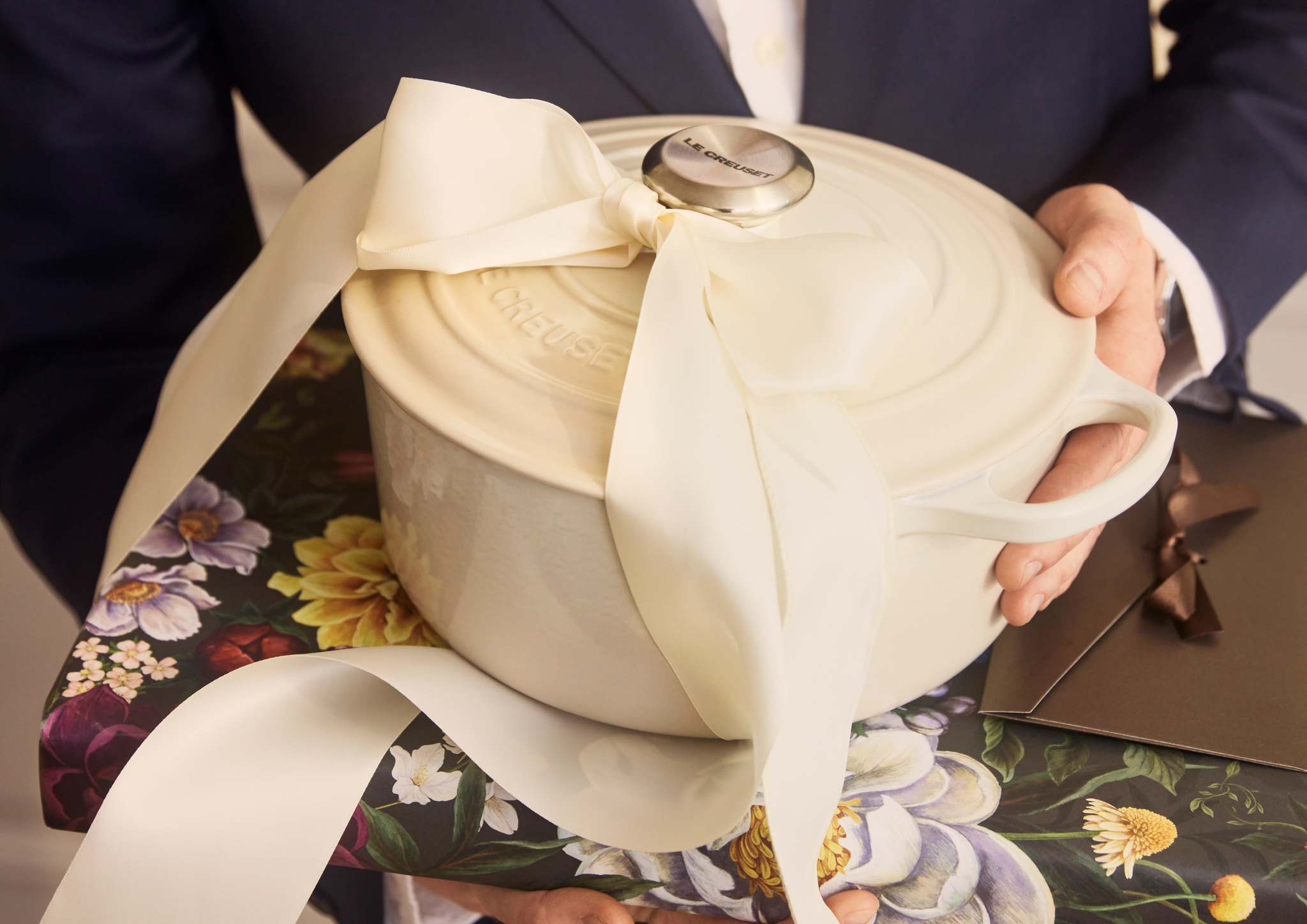 Person i mörk kostym håller en vit gjutjärnsgryta med stor rosett ovanpå ett blommigt inslaget paket, redo att ges som present.
