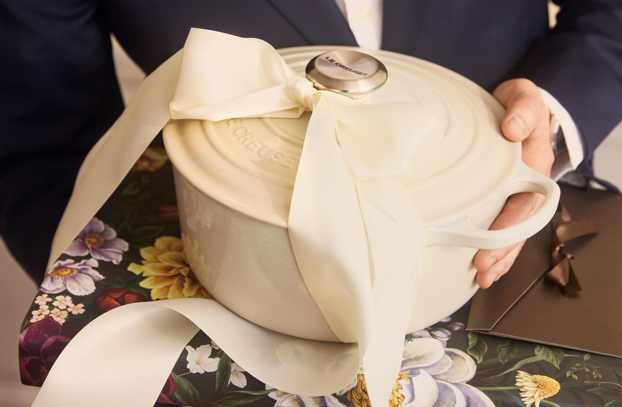 Person i elegant kostym håller en vit gjutjärnsgryta med stor rosett ovanpå ett blommigt inslaget paket, redo att ges som present.