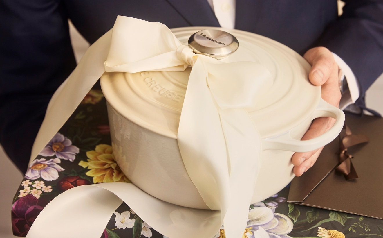 En person i mörk kostym håller en vit gjutjärnsgryta med stor rosett ovanpå ett blommigt inslaget paket.