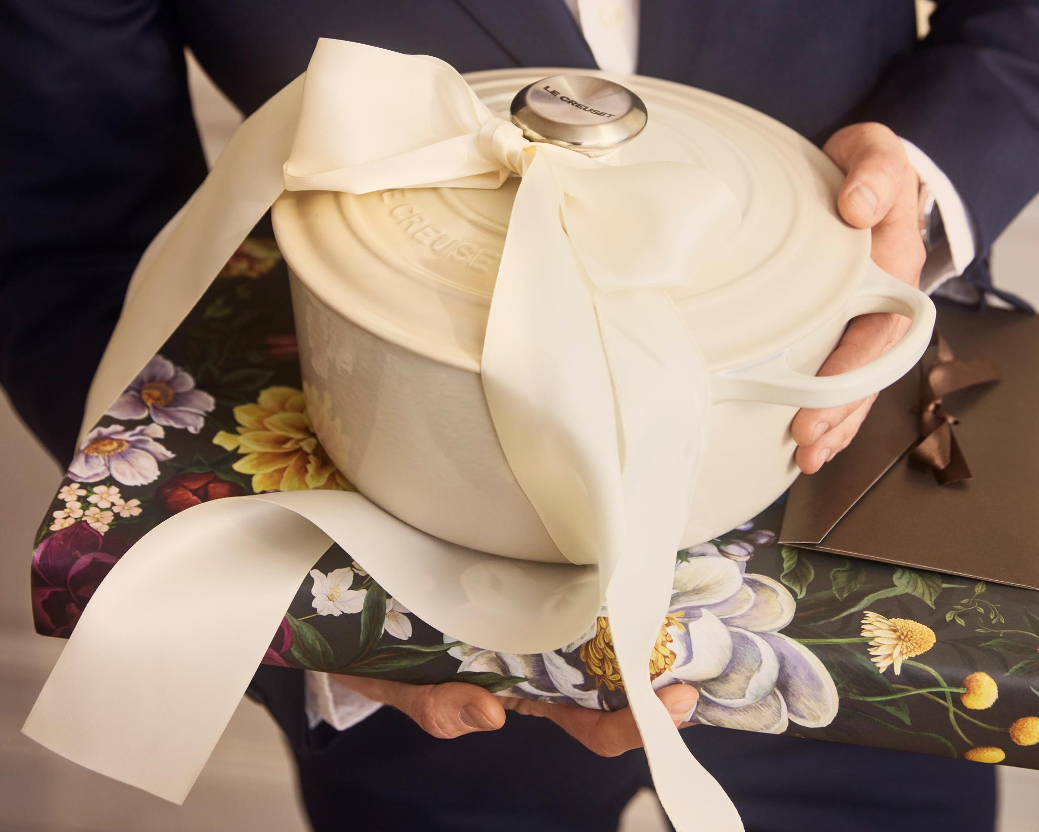 Mann i dress holder en gavepakket Le Creuset-gryte med stor kremfarget sløyfe.