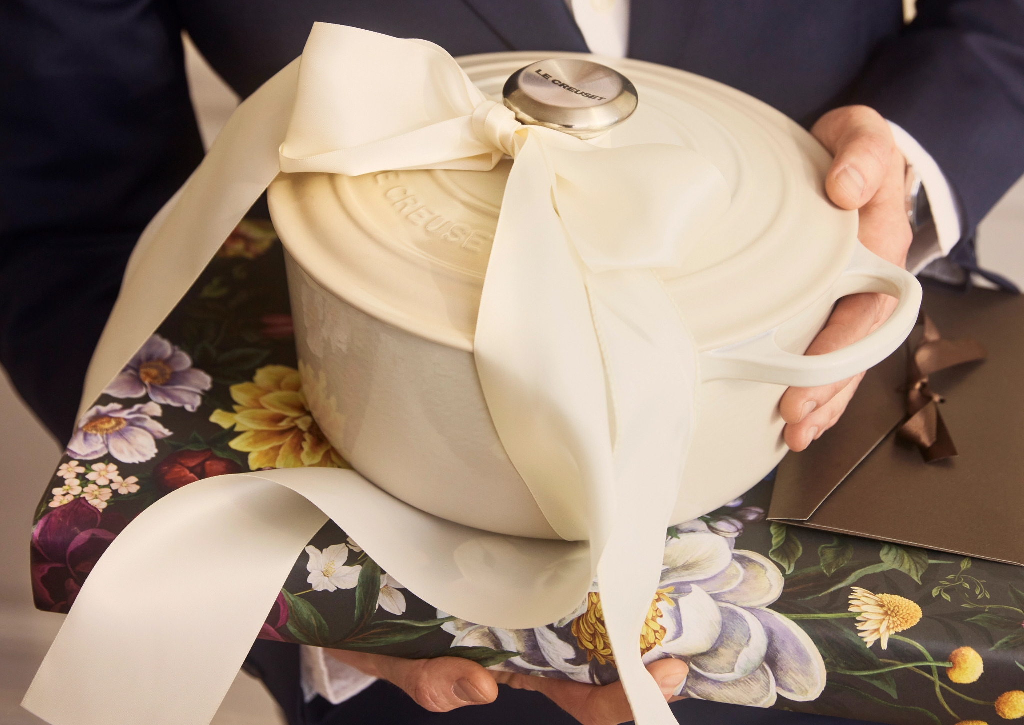 En person i dress holder en kremfarget Le Creuset-gryte med stor sløyfe oppå en blomstrete gaveeske.