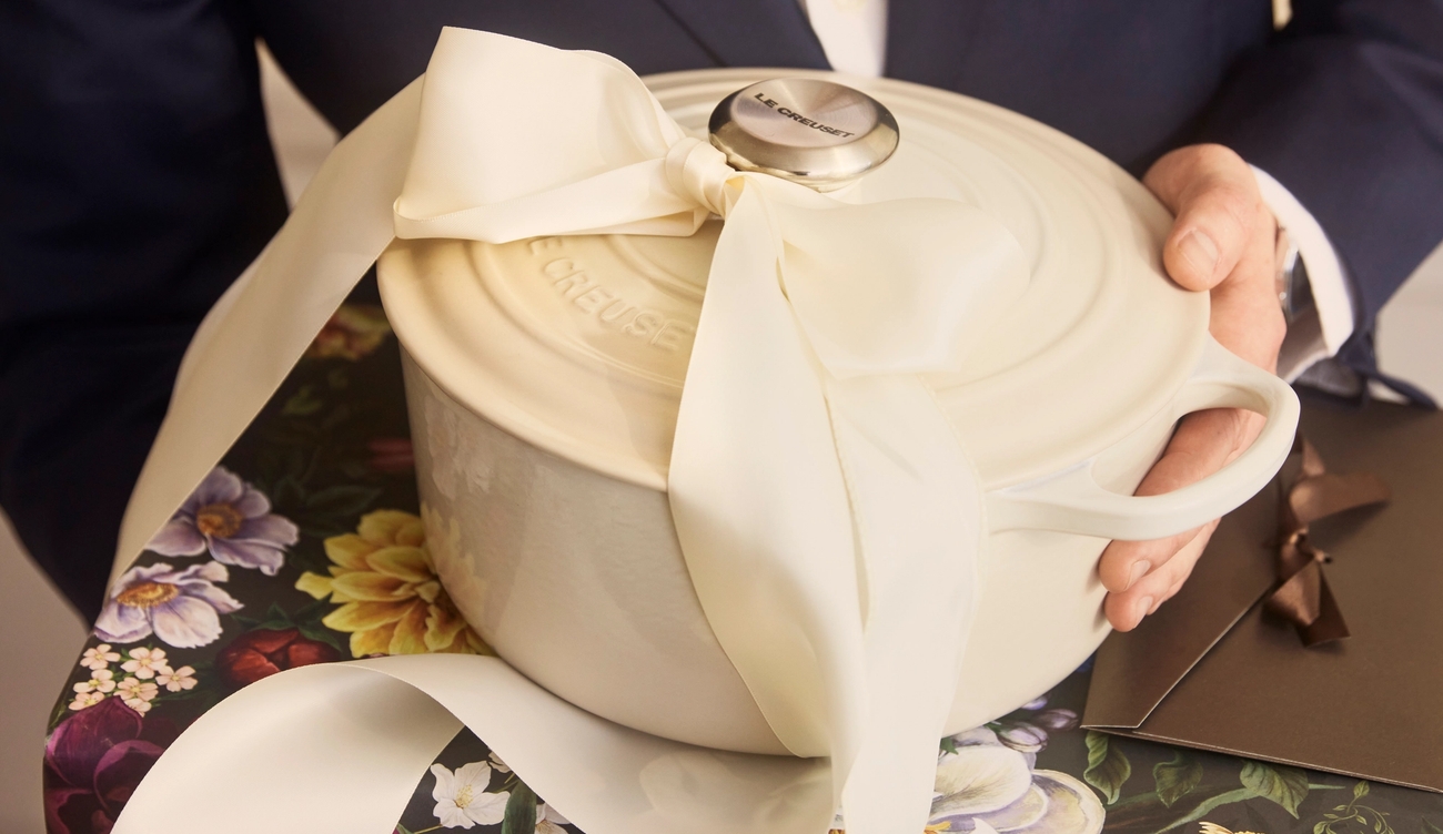 En person i kostym håller en vit Le Creuset-gryta med stor rosett ovanpå ett blommigt inslaget paket, redo att ges som present.