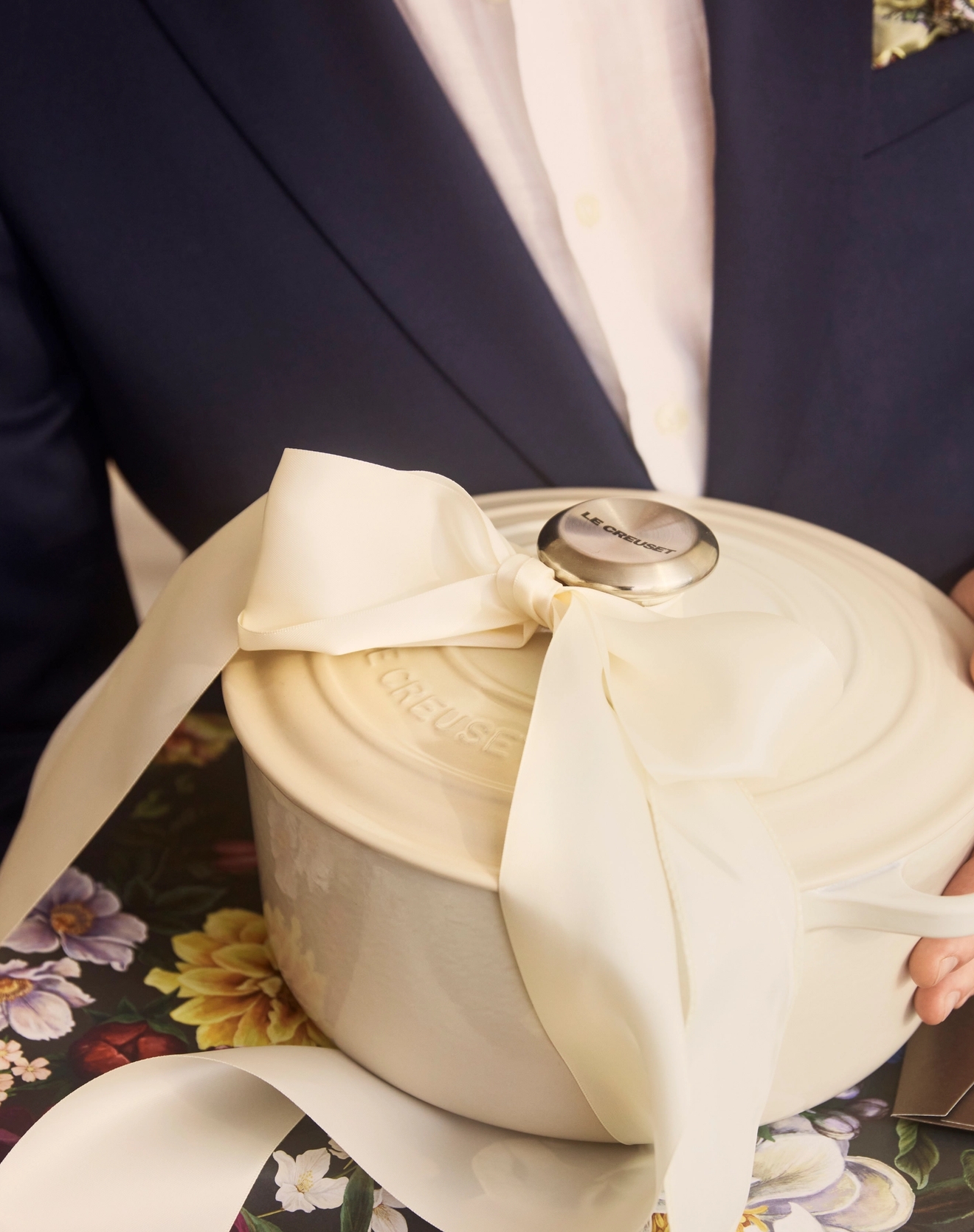 Mann i dress holder en kremfarget Le Creuset-gryte med stor sløyfe oppå en blomstrete gaveeske.